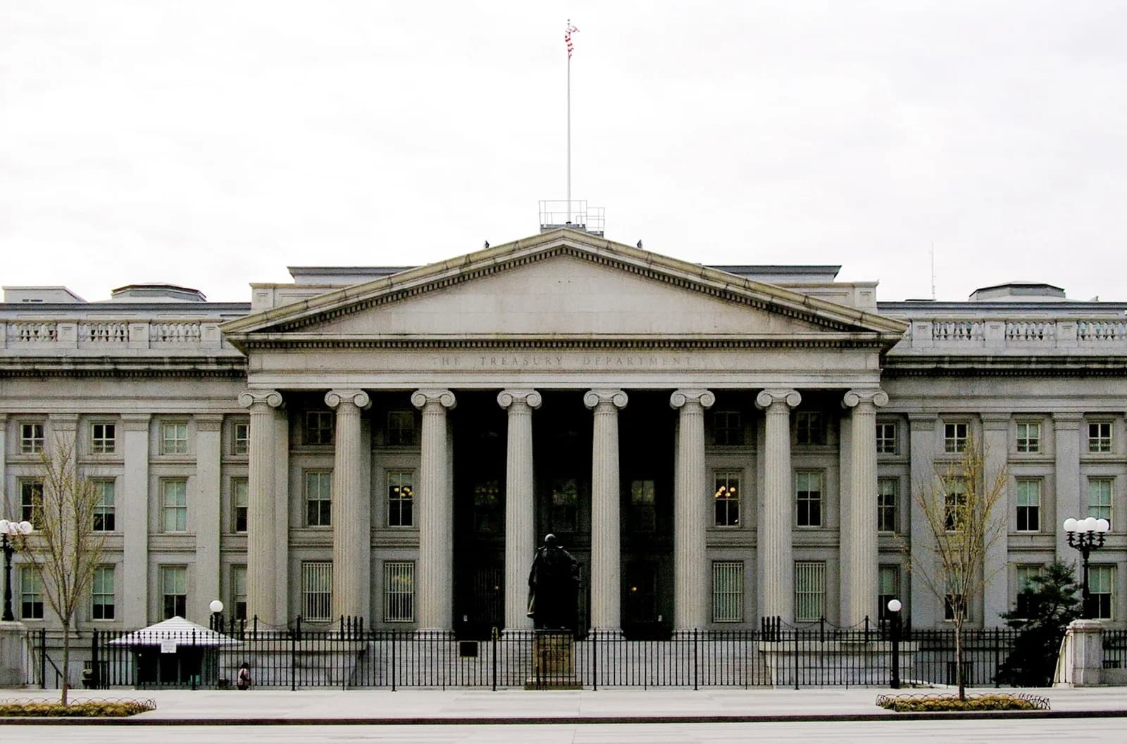A large, neoclassical stone building with a prominent portico featuring tall columns and a triangular pediment. A statue stands in front of the building, behind a black metal fence.