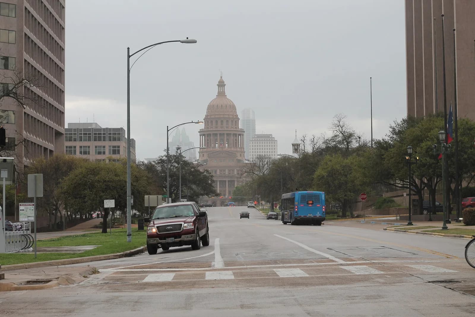 A wide street leads towards a large domed building in the distance under an overcast sky. Cars and a bus are visible on the road, with other buildings and trees lining the sides.