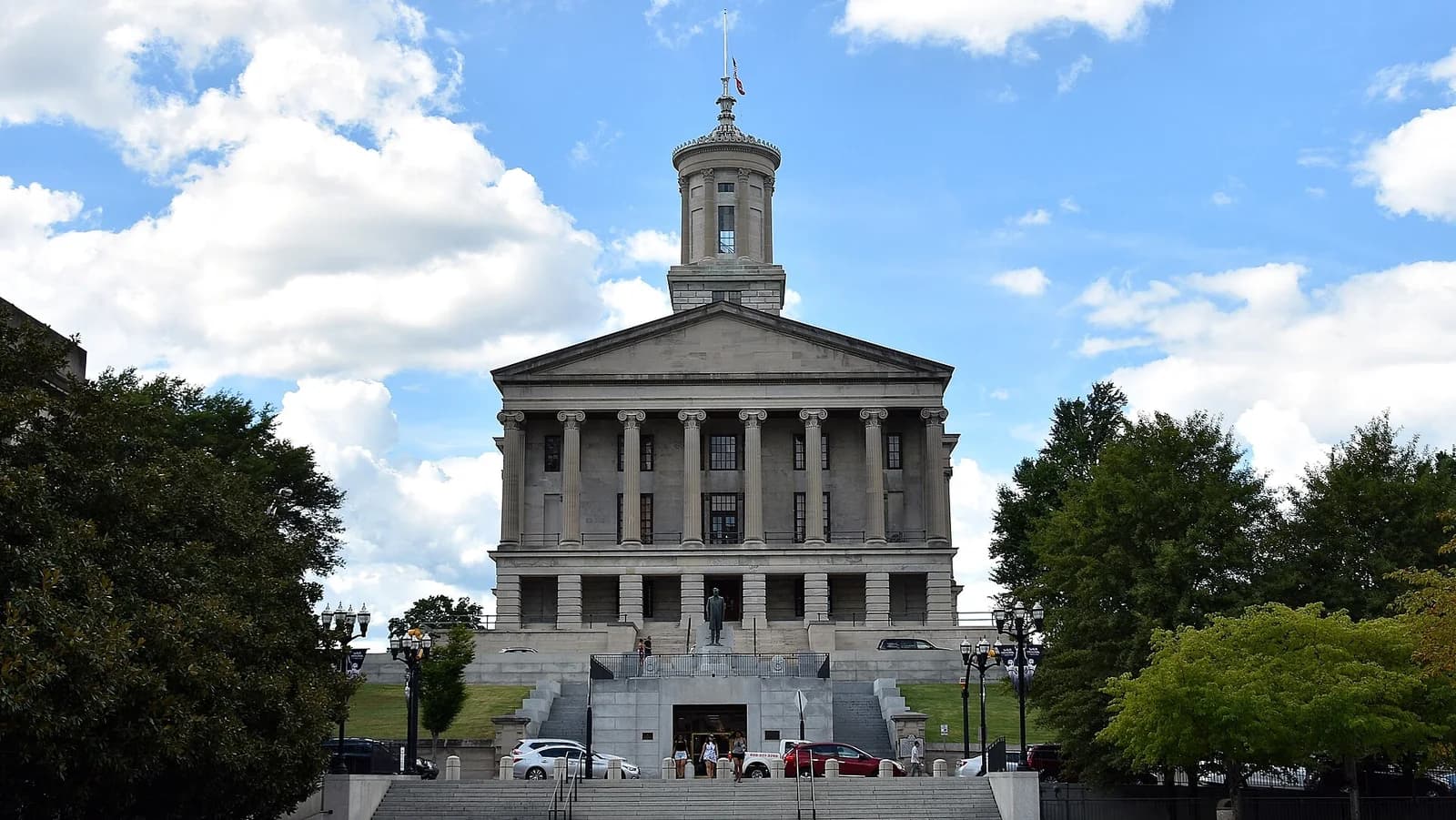 A large, light-colored stone building with a classical facade and a domed cupola on top stands under a blue sky with white clouds. A wide set of stairs leads up to the building's entrance, with trees and streetlights visible on either side.