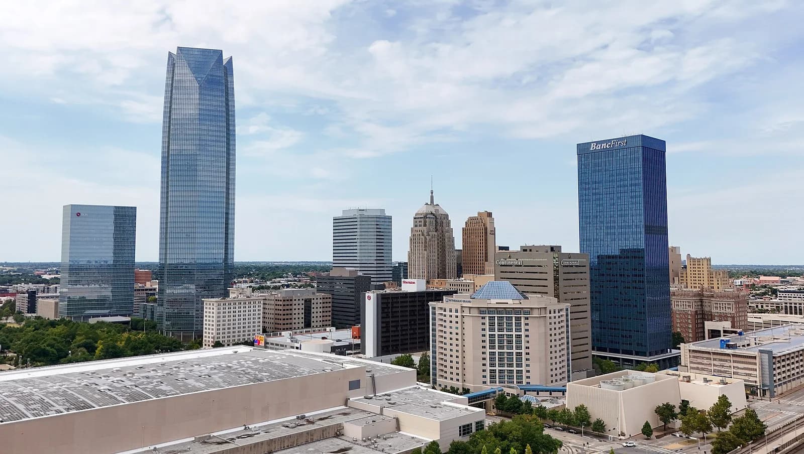 An aerial view of a city skyline shows a mix of modern glass skyscrapers and older, more ornate buildings under a partly cloudy sky. A large, light-colored building with a flat roof is visible in the foreground.