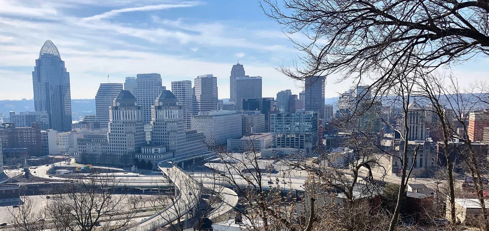A wide view of a city skyline with numerous buildings under a partly cloudy sky, seen from a hillside with bare tree branches in the foreground. Highways and urban infrastructure are visible in the midground leading to the city.