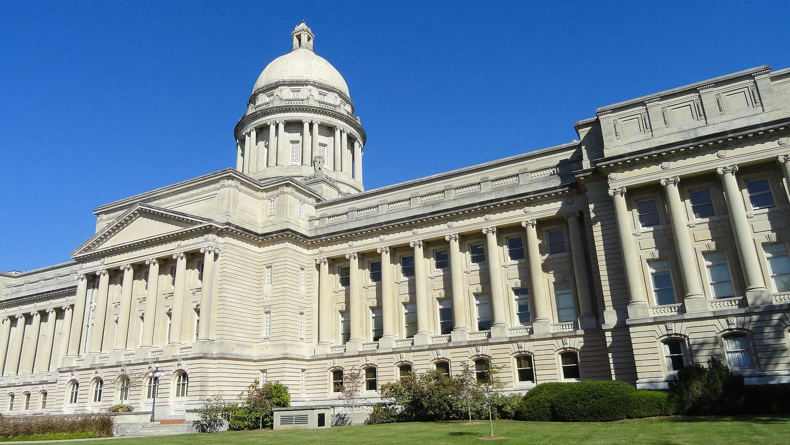 A large, ornate stone building with a prominent dome and columns stands under a clear blue sky. The building is surrounded by green grass and some bushes.