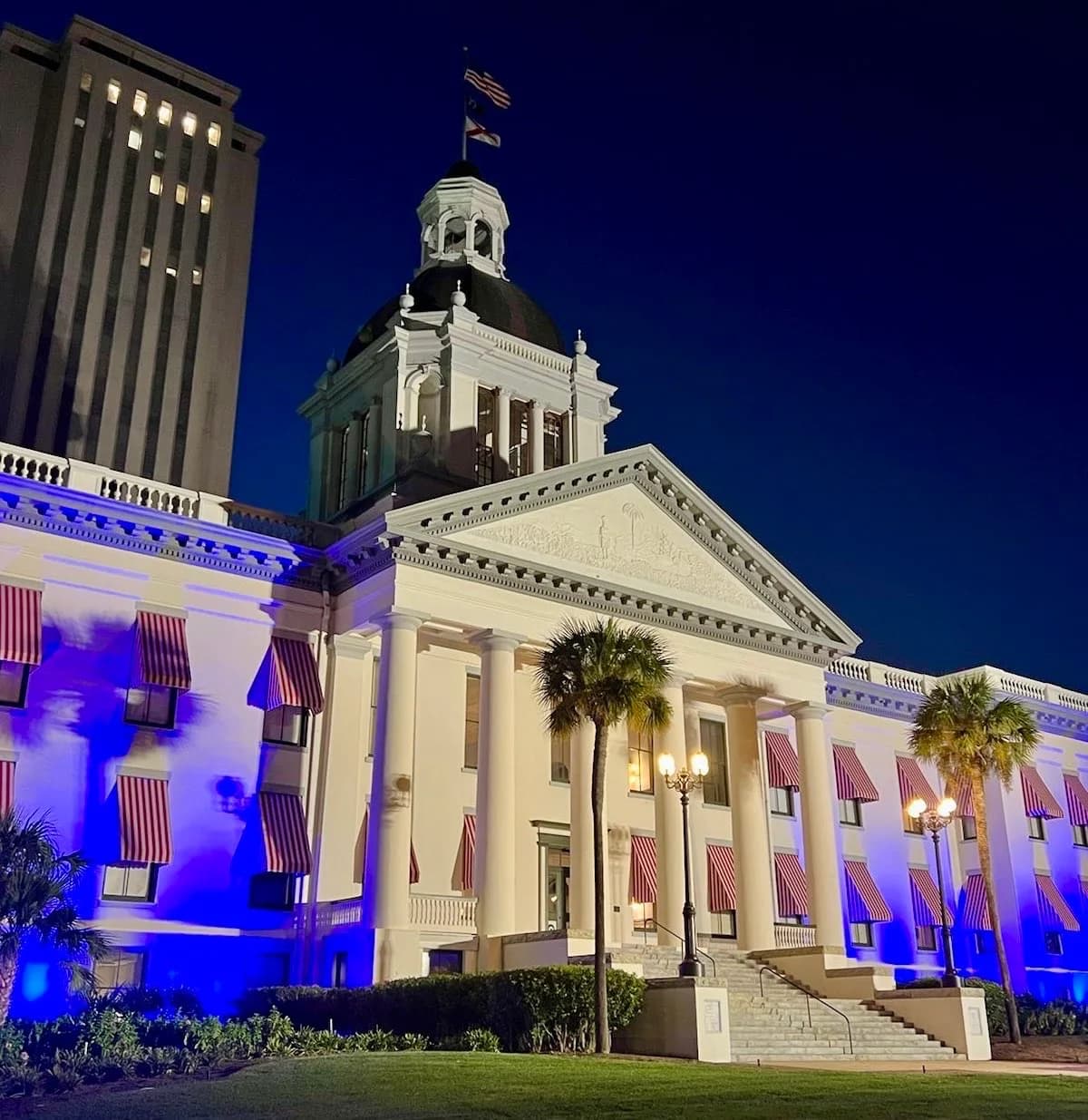 A grand white building with a dome and columns is illuminated with blue lights at night. Red and white striped awnings adorn the windows, and a tall modern building stands behind it against a dark blue sky.