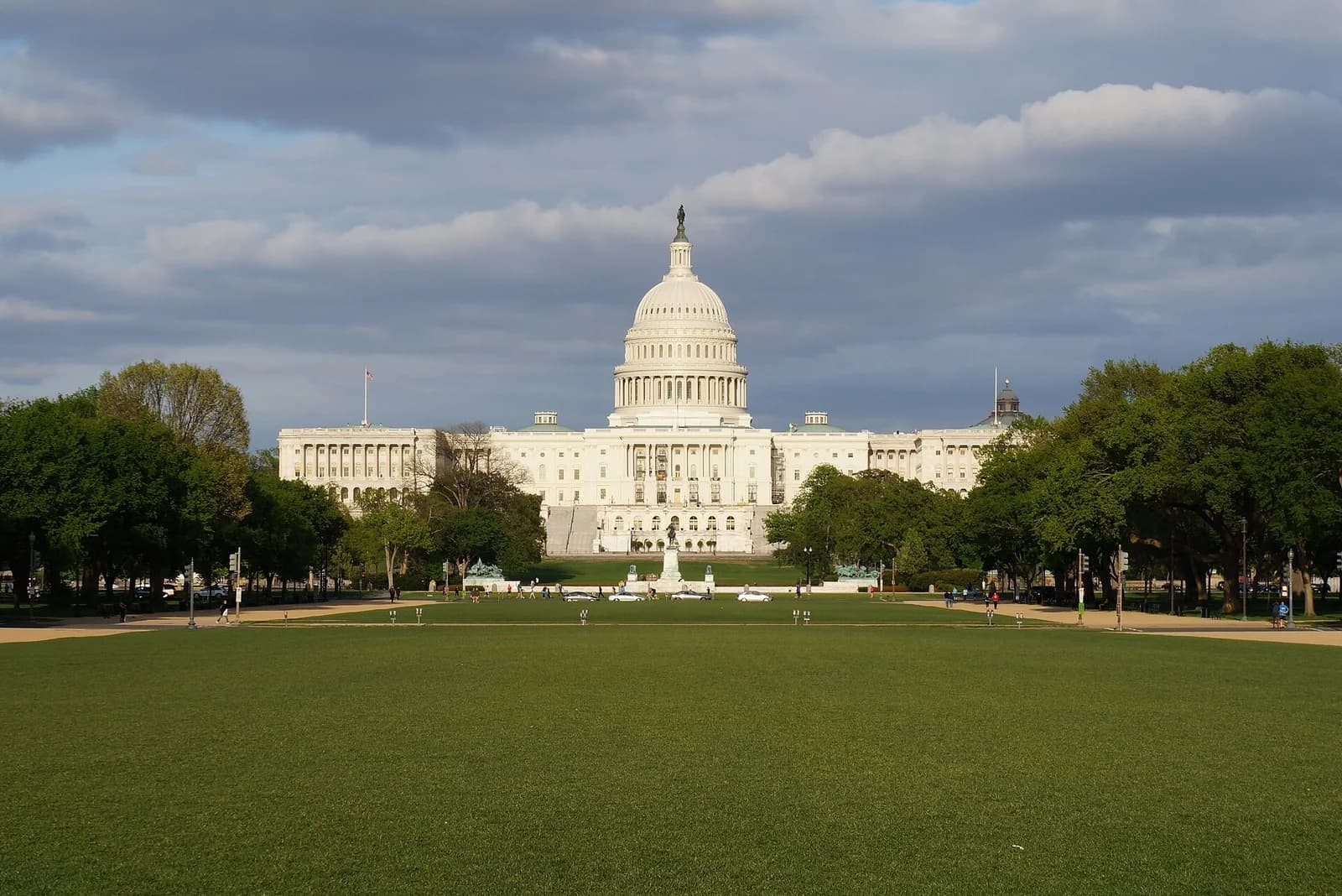 A large white domed building with columns stands prominently at the end of a wide green lawn under a partly cloudy sky. Trees line the sides of the lawn, and a few distant people are visible.