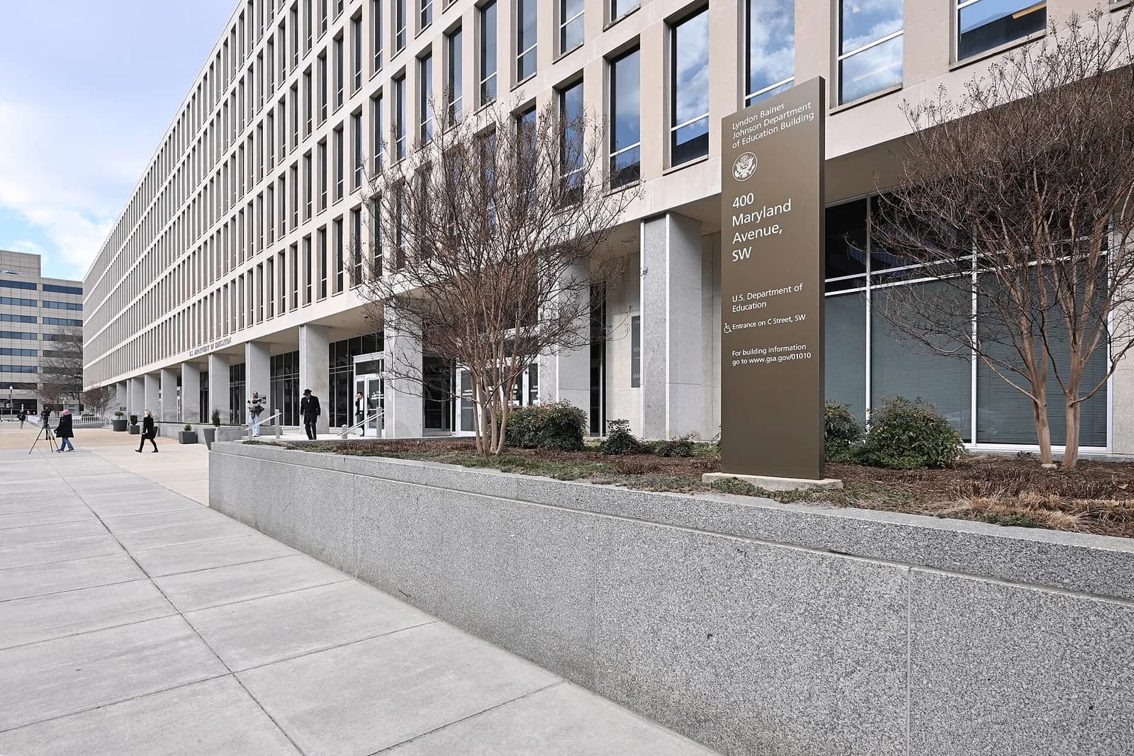 A long, modern institutional building with many windows and a prominent sign stands beside a paved walkway. Several people are visible walking near the building's entrance.