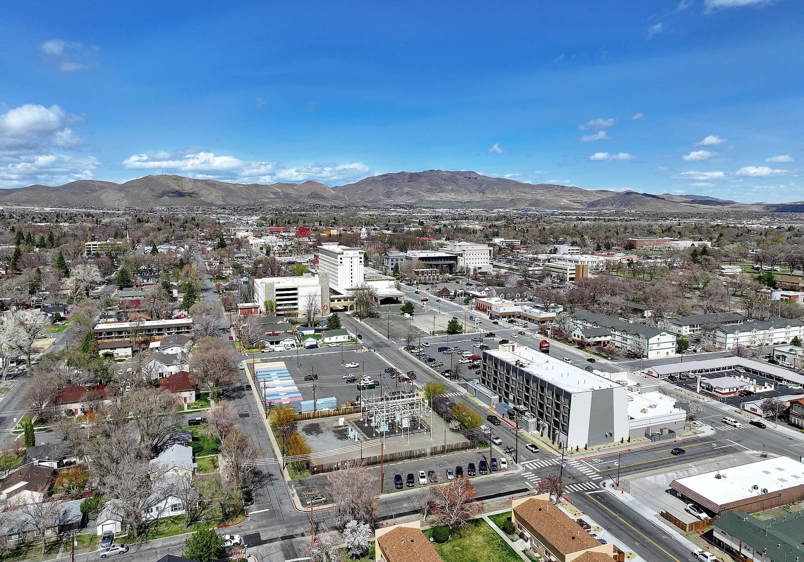 An aerial view of a city shows a mix of residential and commercial buildings, with a prominent electrical substation in the foreground. Mountains rise in the distance under a bright blue sky with scattered clouds.