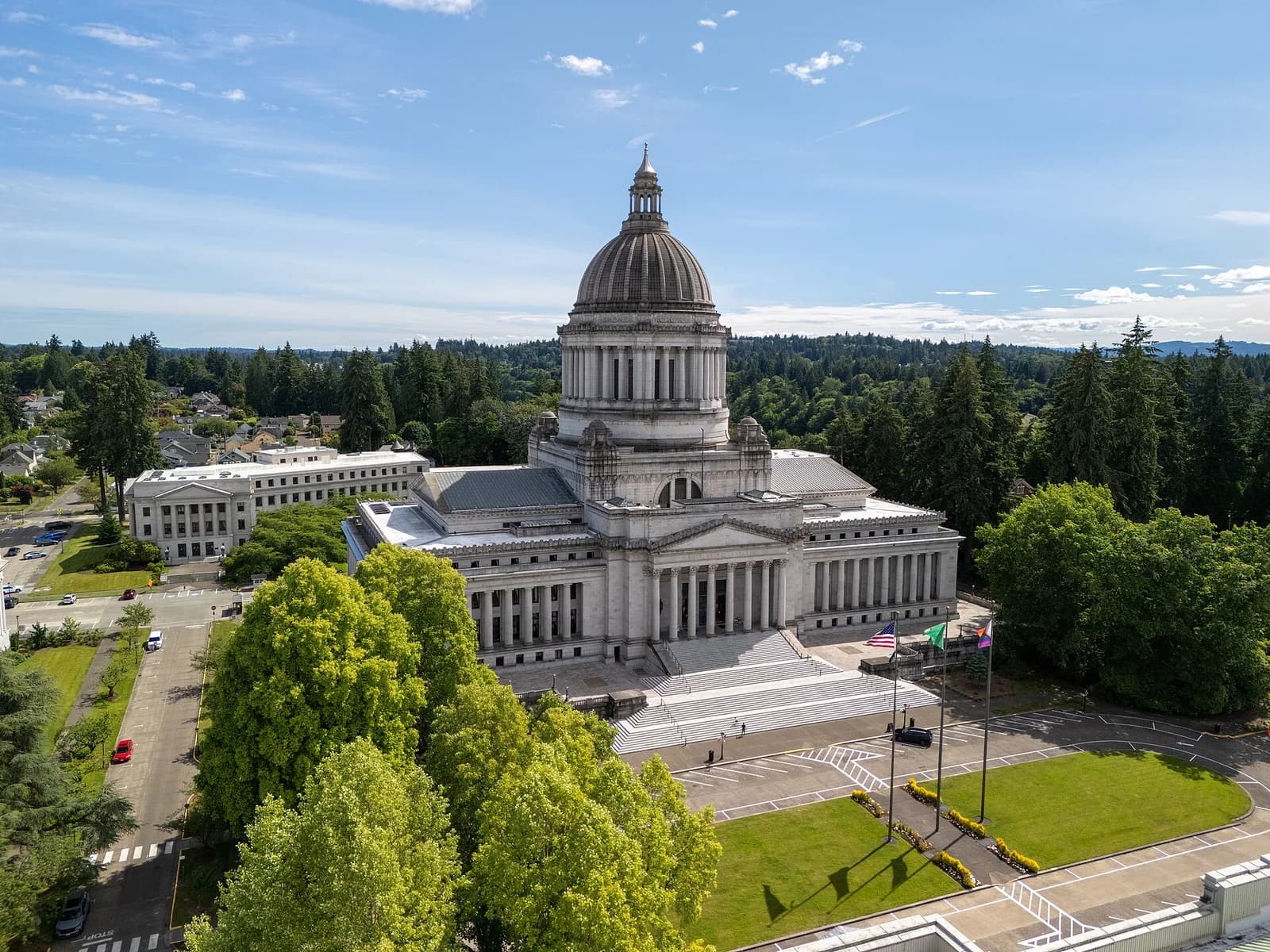 An aerial view shows a large white domed building with columns and steps, surrounded by green trees and a paved area with flags. In the background, more buildings and a dense forest are visible under a partly cloudy sky.