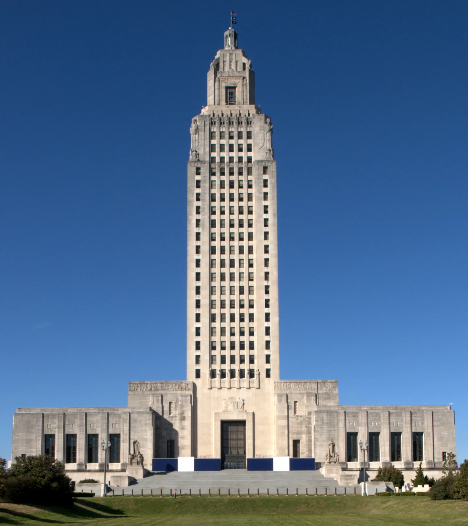 A tall, light-colored Art Deco skyscraper with a stepped-back top rises from a wider, lower base. The building is set against a clear blue sky, with green grass and some trees in the foreground.