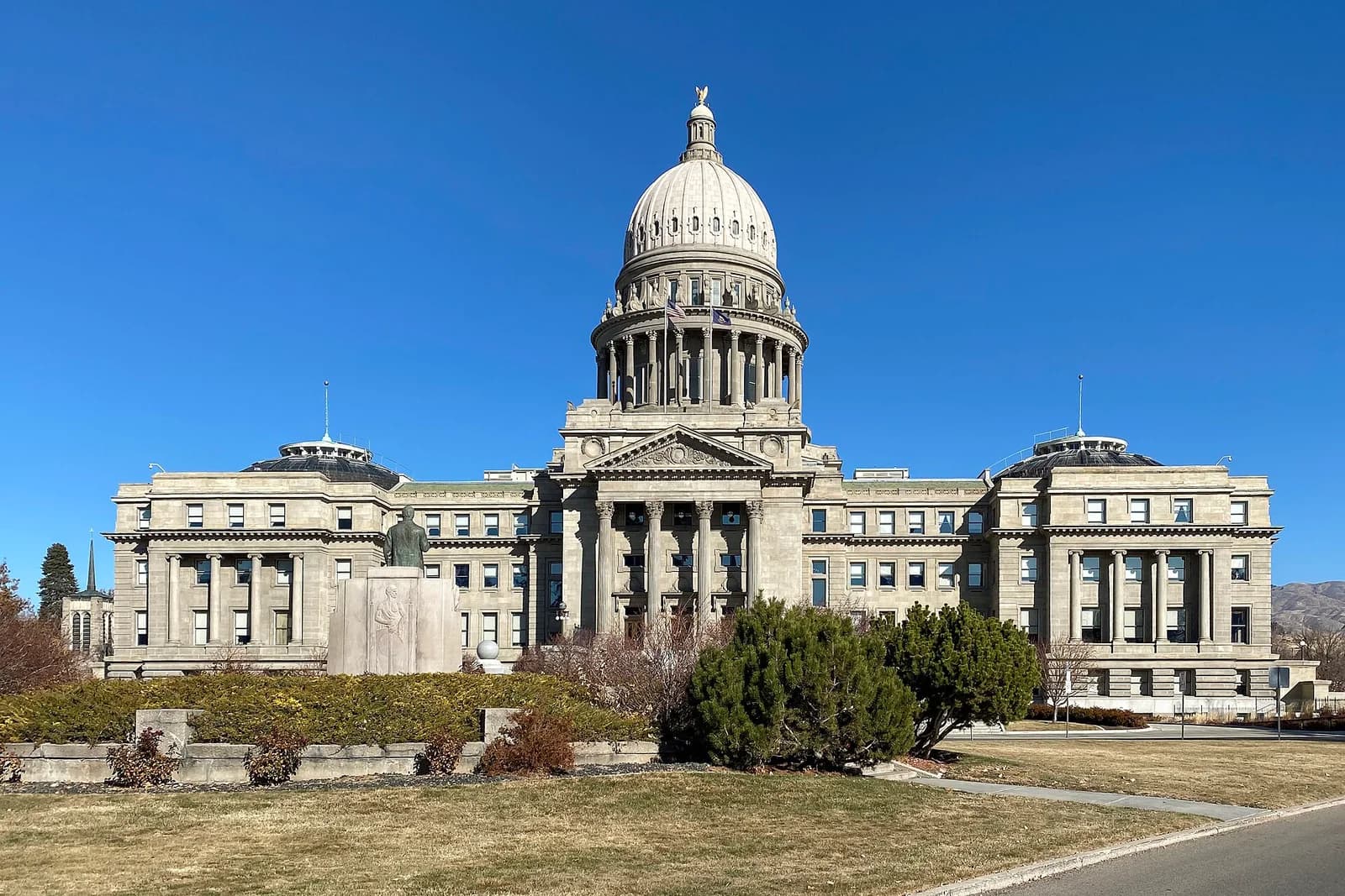 A grand, light-colored stone building with a large central dome and columns stands under a clear blue sky. In the foreground, there is a grassy area with some bushes and a statue.