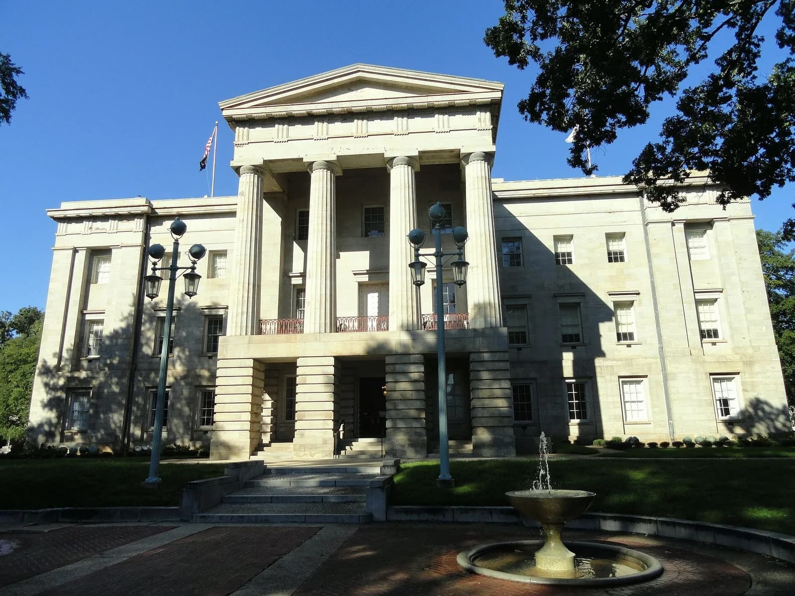 A large, light-colored stone building with a prominent portico featuring tall columns and a triangular pediment stands under a clear blue sky. In the foreground, a small fountain with water spraying upwards is visible, along with brick paving and green grass.