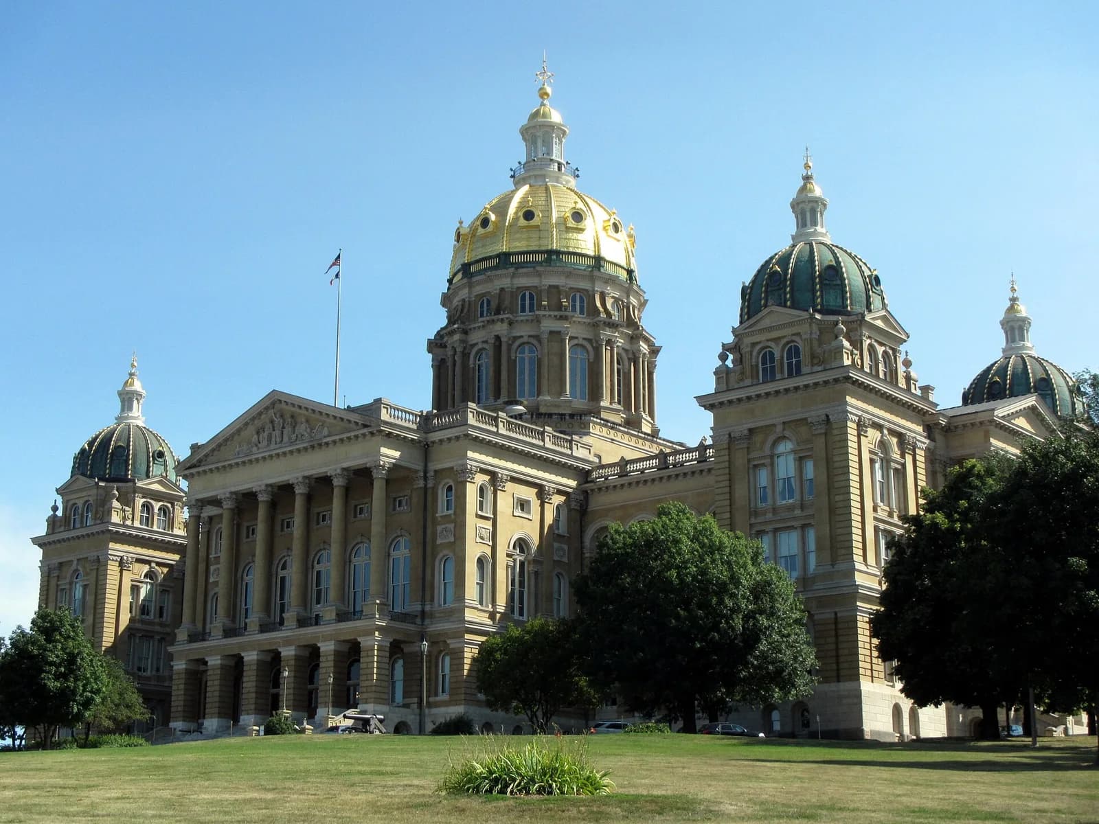 A large, ornate building with a prominent golden dome and several smaller green domes stands on a grassy hill under a clear blue sky. The building features classical architectural elements including columns and pediments.