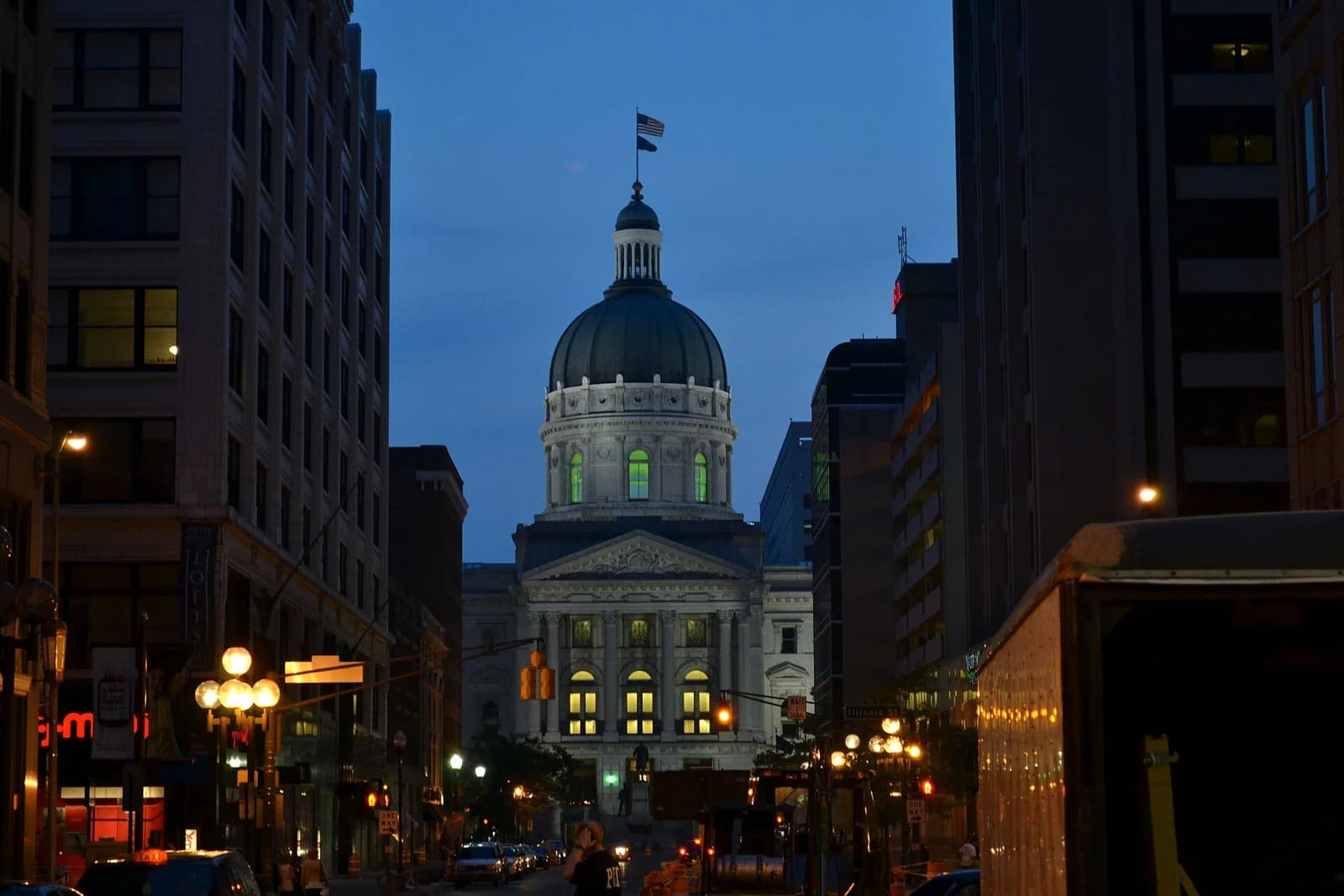 A grand domed building with a flag on top is visible at the end of a city street at dusk, flanked by taller, darker buildings. Streetlights illuminate the road and some cars, with a large truck partially visible on the right.