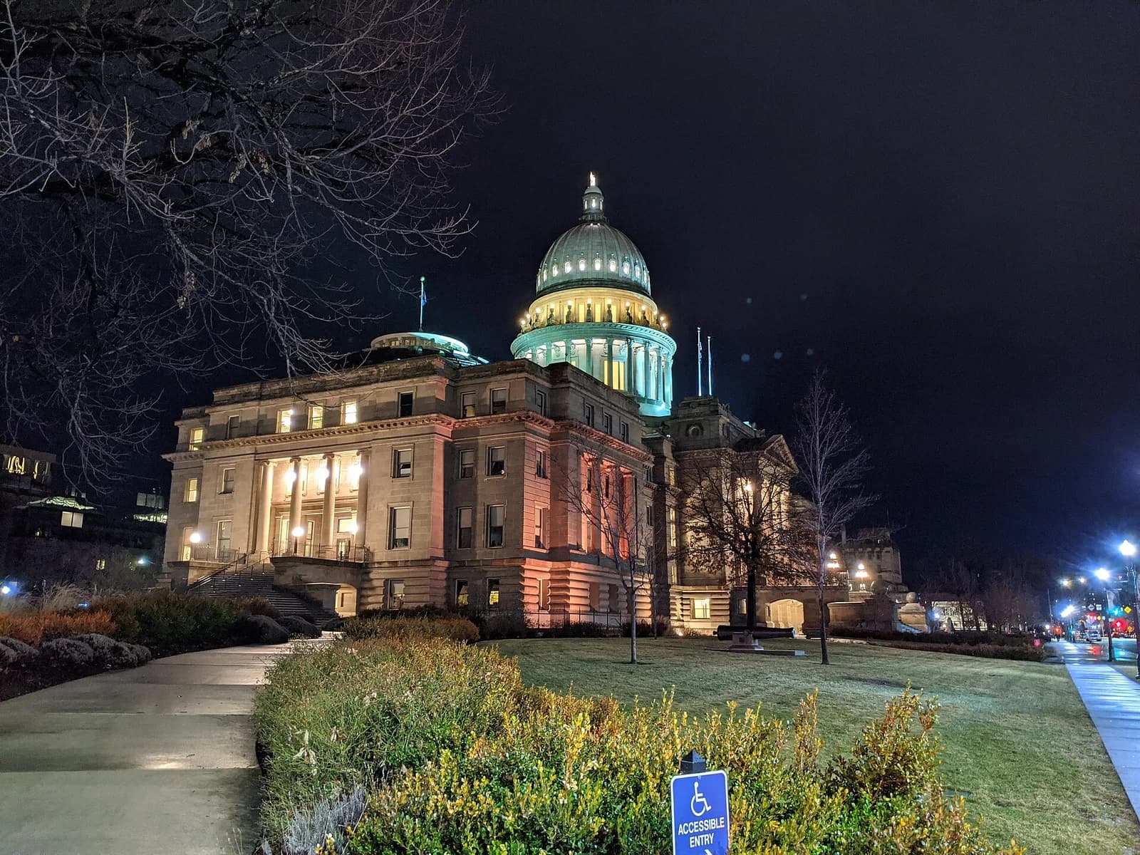 A grand, illuminated building with a large dome is visible at night, surrounded by bare trees and manicured bushes. A paved path leads towards the building, and a blue sign for "Accessible Entry" is in the foreground.