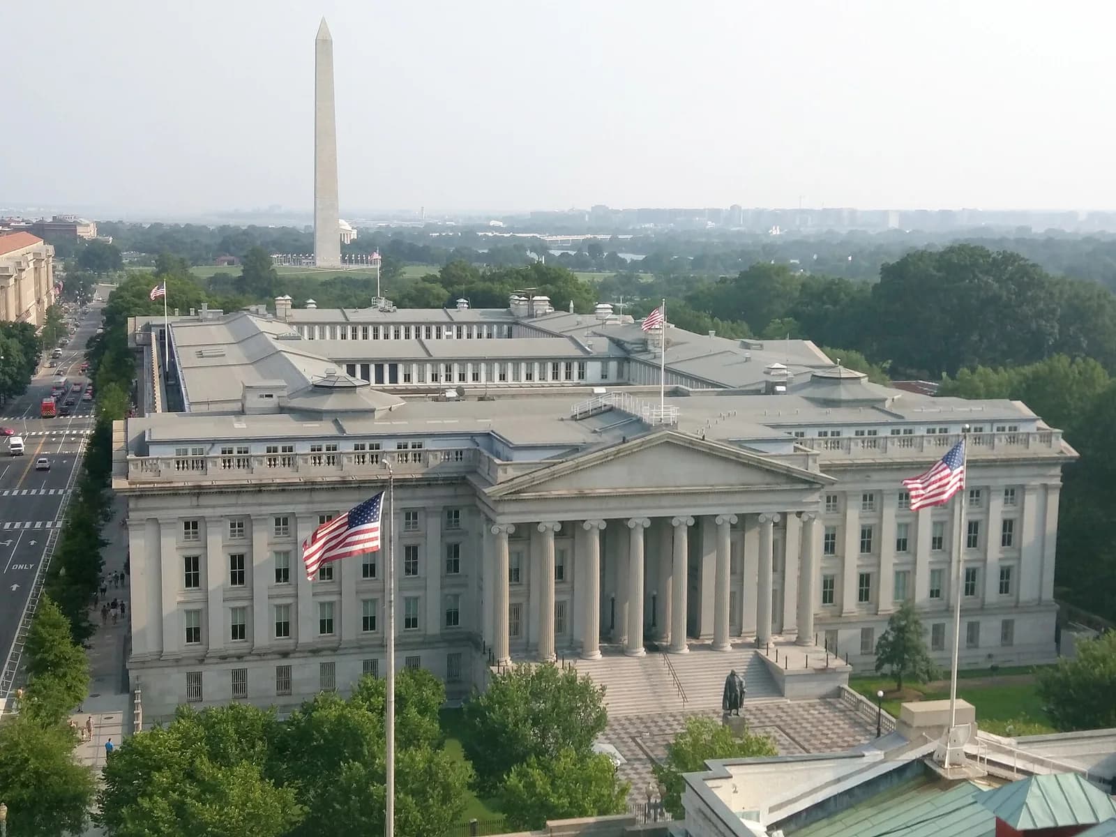 An aerial view shows a large, light-colored neoclassical building with a prominent portico and columns, flanked by American flags. In the background, a tall, slender obelisk rises above a tree-lined landscape, with a hazy city skyline visible in the distance.