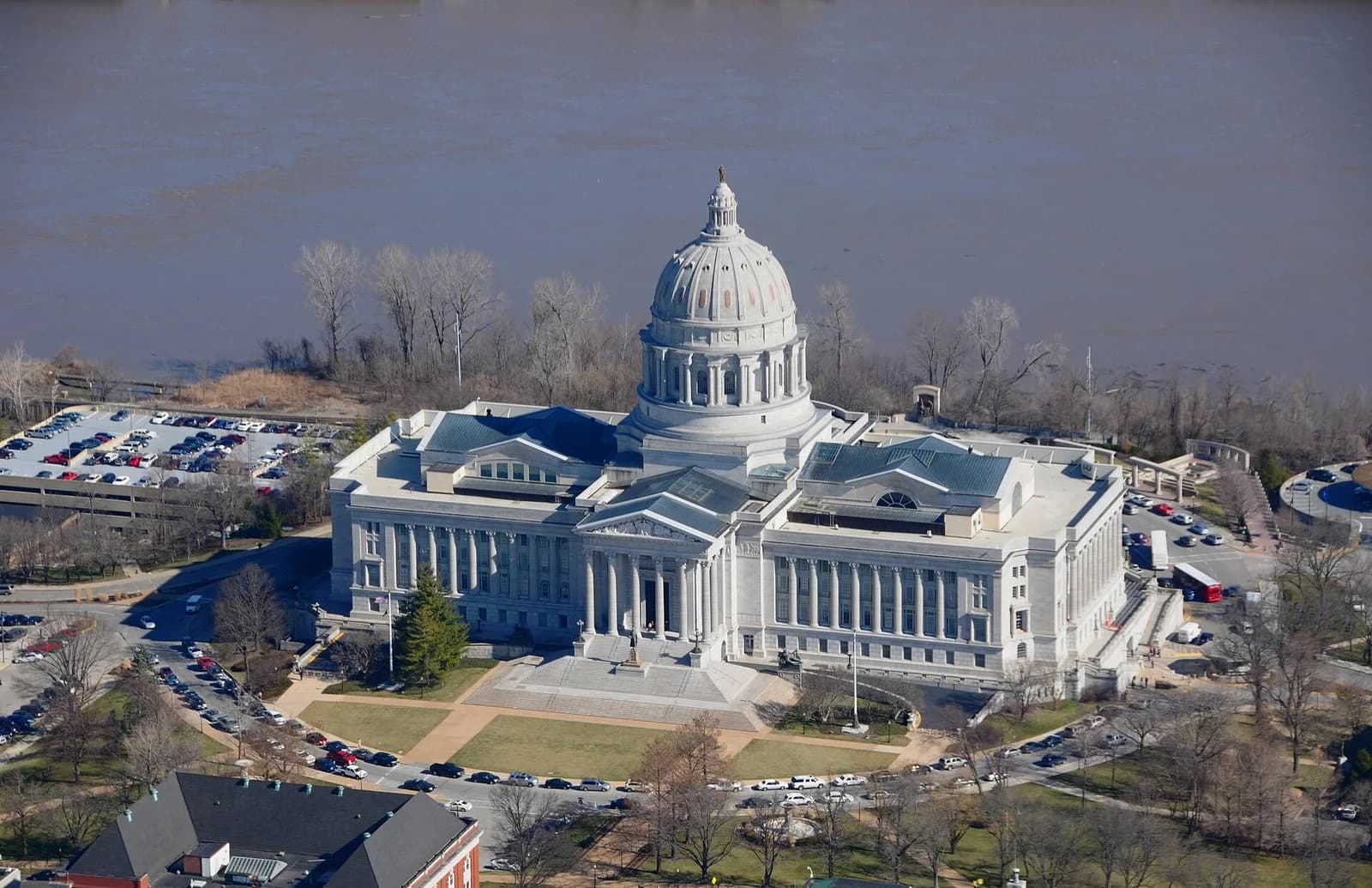 An aerial view shows a large, light-colored domed building with columns, surrounded by green lawns, roads with parked cars, and a river in the background. Bare trees line the riverbank.