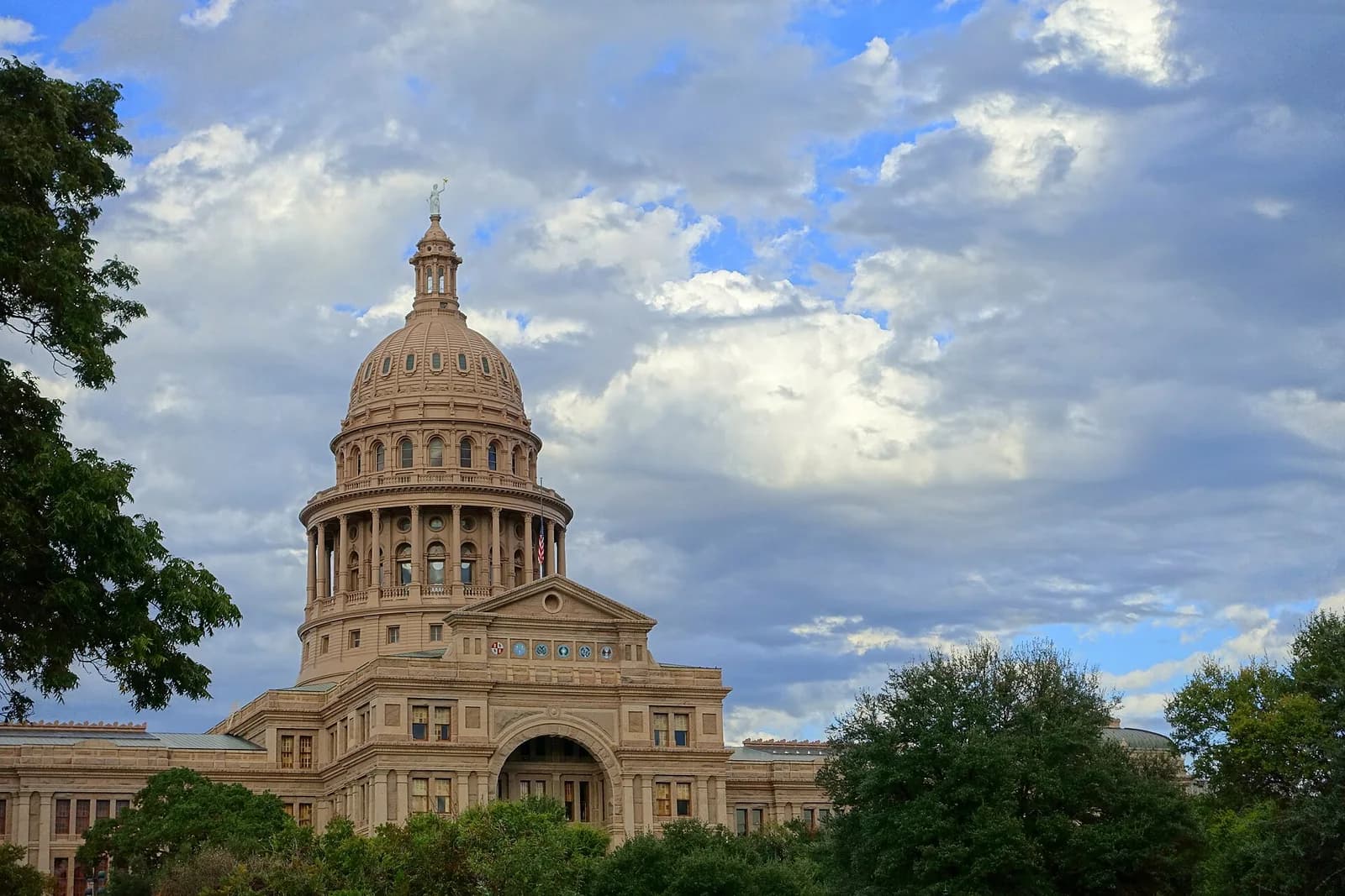 A large, light-colored stone building with a prominent dome and cupola stands under a dramatic sky with white and gray clouds. Green trees frame the bottom and sides of the building.