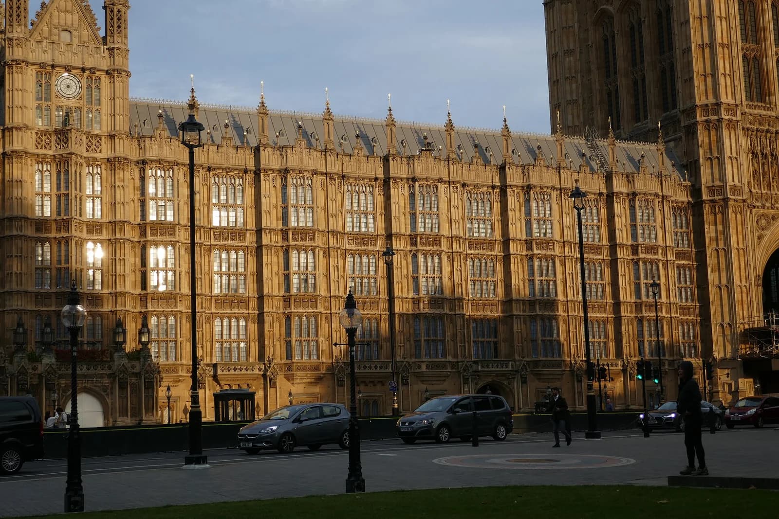A large, ornate stone building with many windows and spires is illuminated by sunlight. Cars and a few pedestrians are visible on a street in front of the building.
