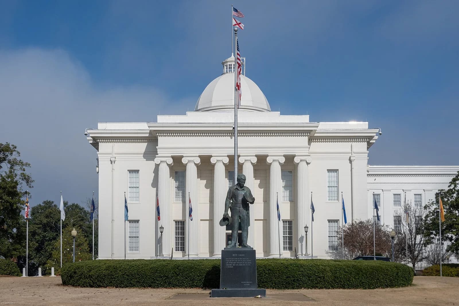 A white neoclassical building with a large dome and columns stands under a blue sky. In front of the building is a statue of a man on a pedestal, with a flagpole flying two flags directly behind it.