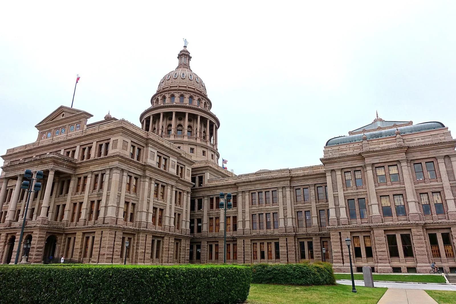 A large, ornate stone building with a prominent dome and a flag flying from its roof is visible under a cloudy sky. Green hedges and grass are in the foreground, with a sidewalk and streetlights also visible.