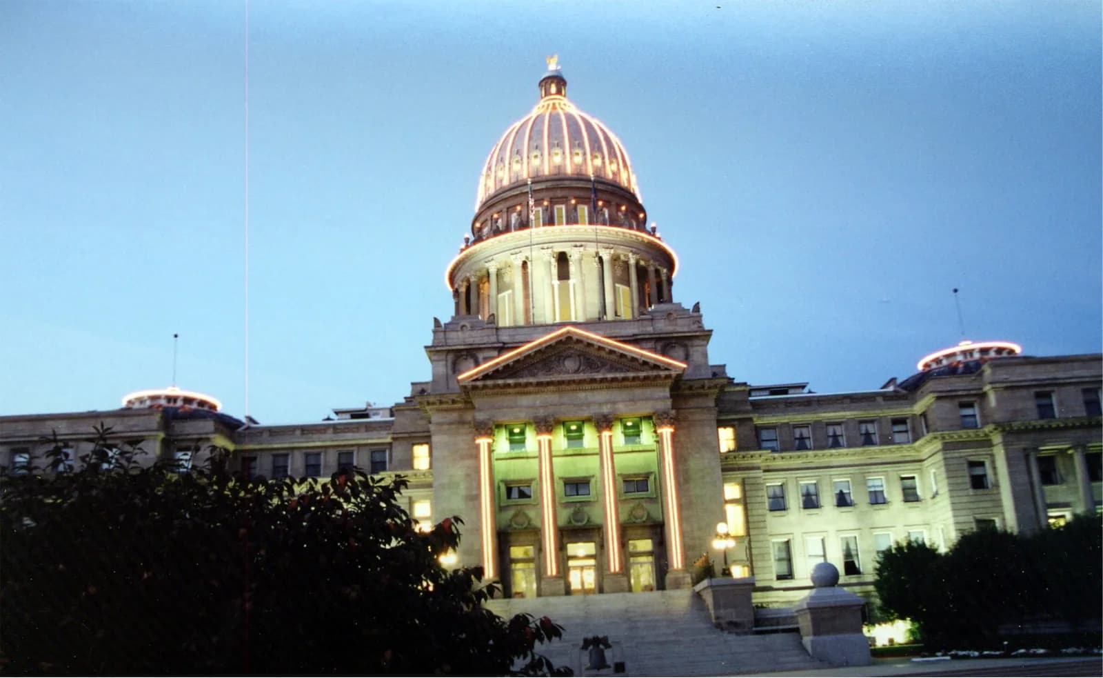 A grand, illuminated building with a large central dome and two smaller domes on either side, seen at dusk or night. The building features classical architecture with columns and is lit with string lights outlining its features.