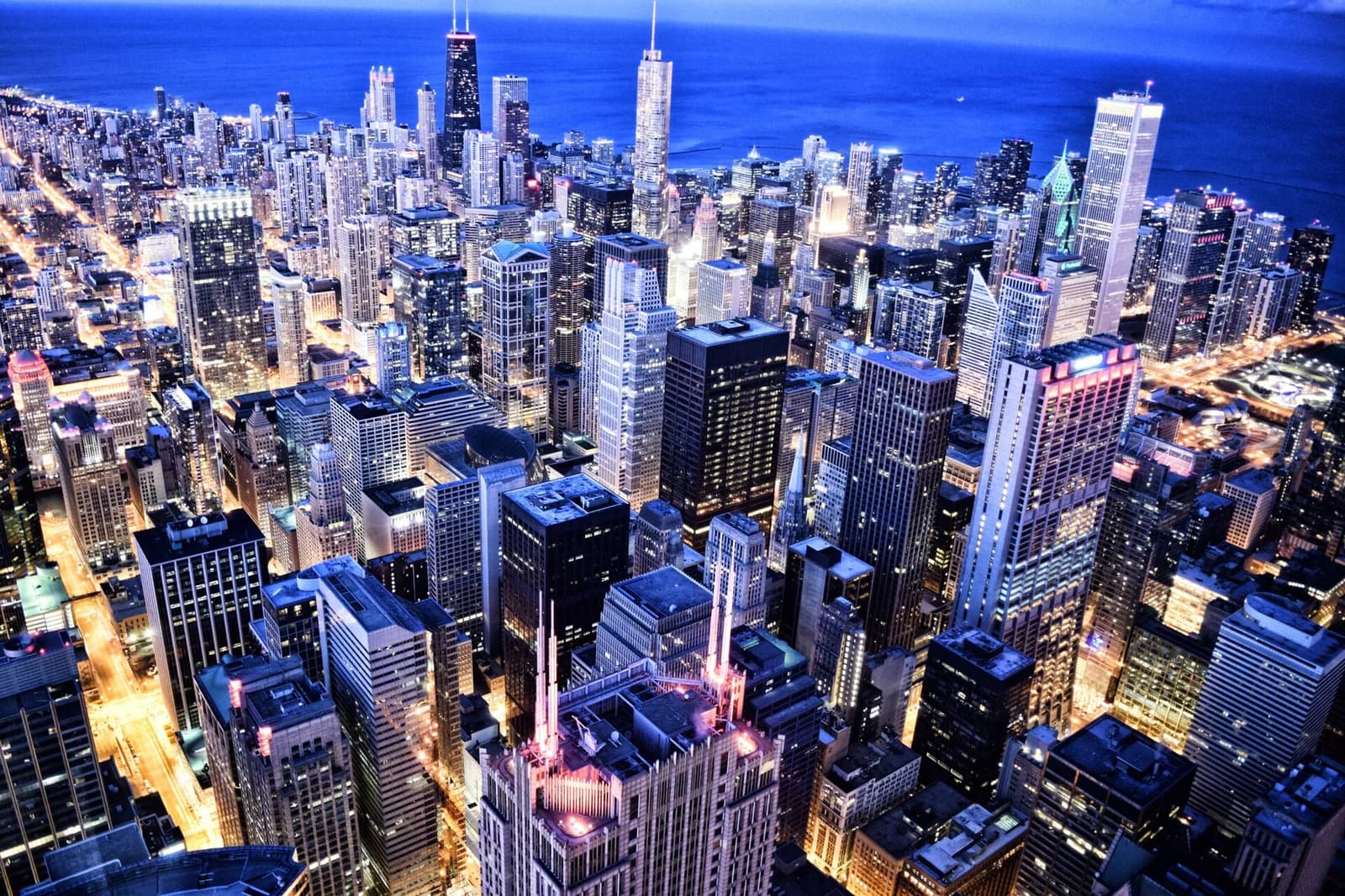 An aerial view of a city at dusk shows numerous skyscrapers and buildings with illuminated windows and streetlights. A large body of water is visible in the background under a dark blue sky.