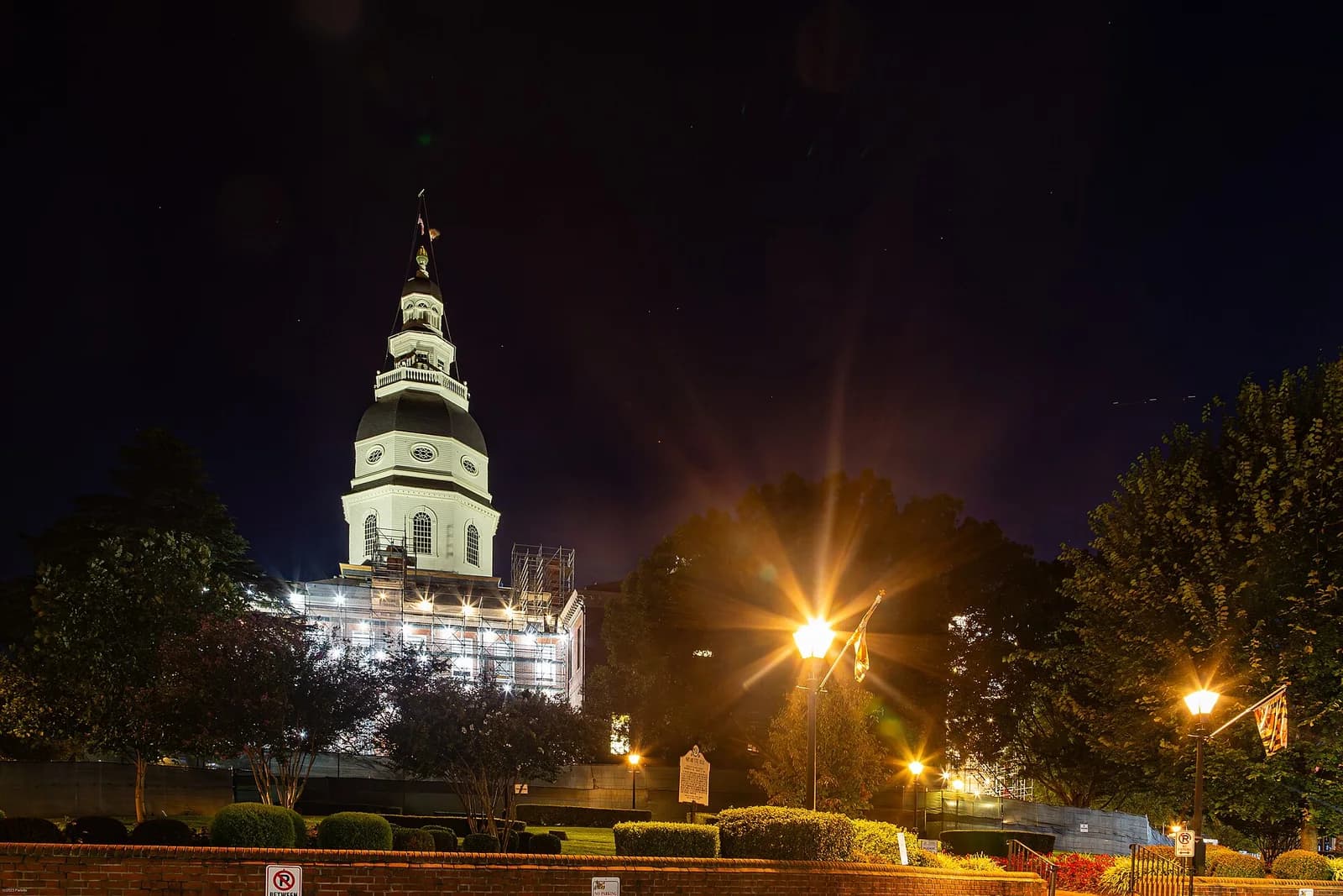 A white domed building with a cupola and spire, partially covered in scaffolding, is illuminated at night. The building is surrounded by trees and streetlights, with a dark sky above.