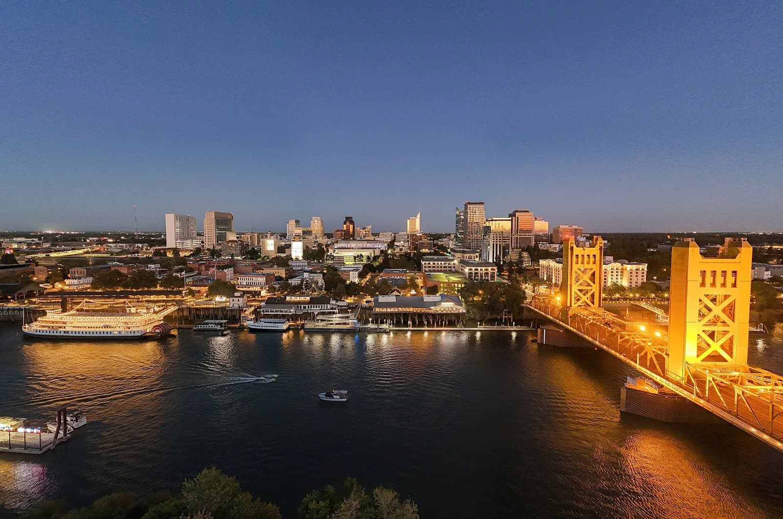 An aerial view of a city skyline at dusk, with a river in the foreground reflecting the lights of buildings and a prominent golden-yellow bridge. Several boats are visible on the water, including a large paddlewheel boat docked near the city.