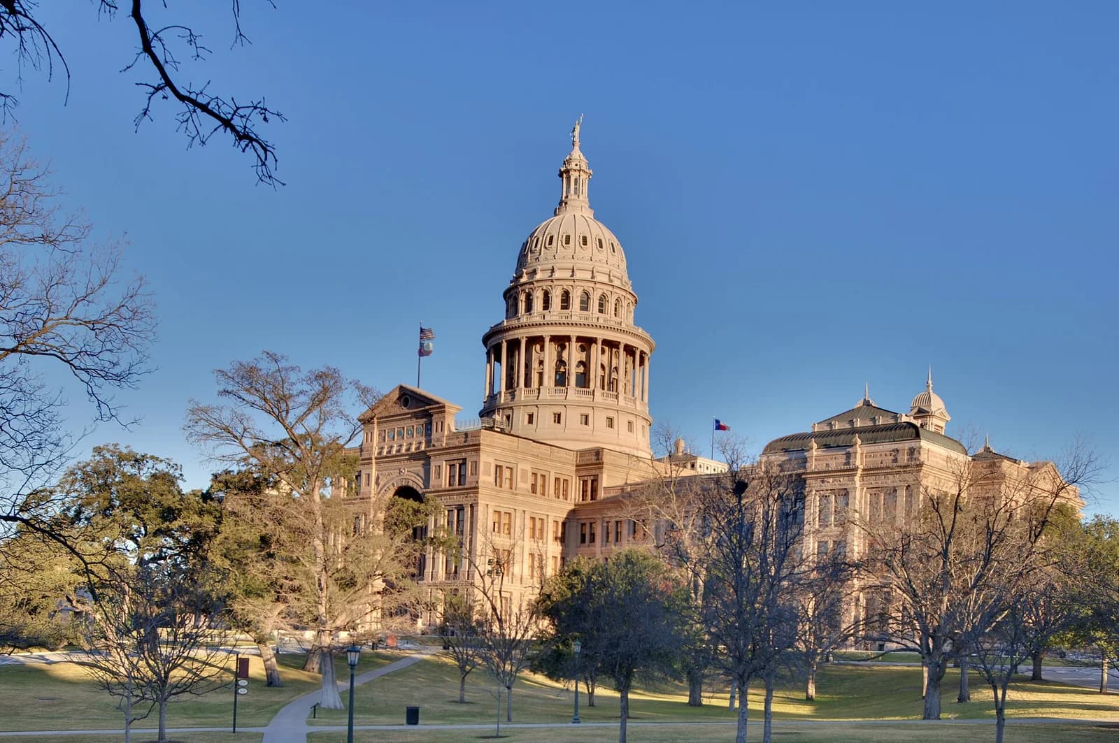 A large, ornate stone building with a prominent dome and cupola stands under a clear blue sky. The building is surrounded by a grassy lawn with bare trees and a paved pathway.