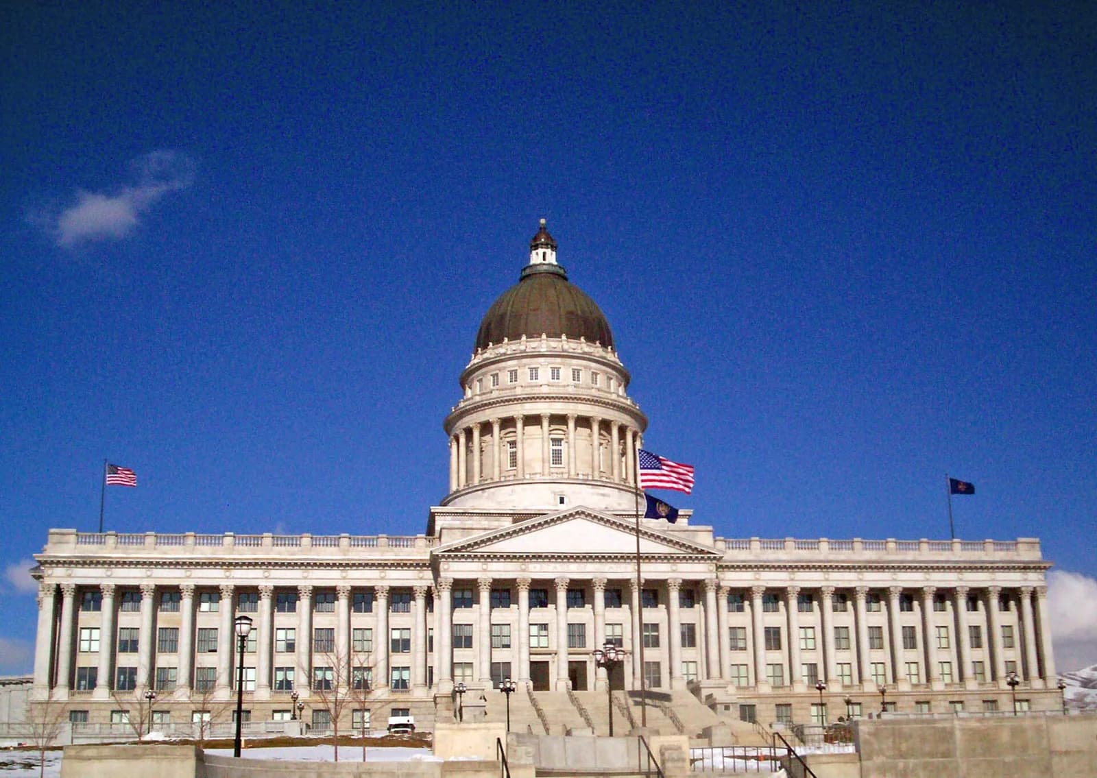 A grand, white neoclassical building with a large dome and columns stands under a clear blue sky. American and other flags fly from poles on the building's facade, with some snow visible on the ground in front.