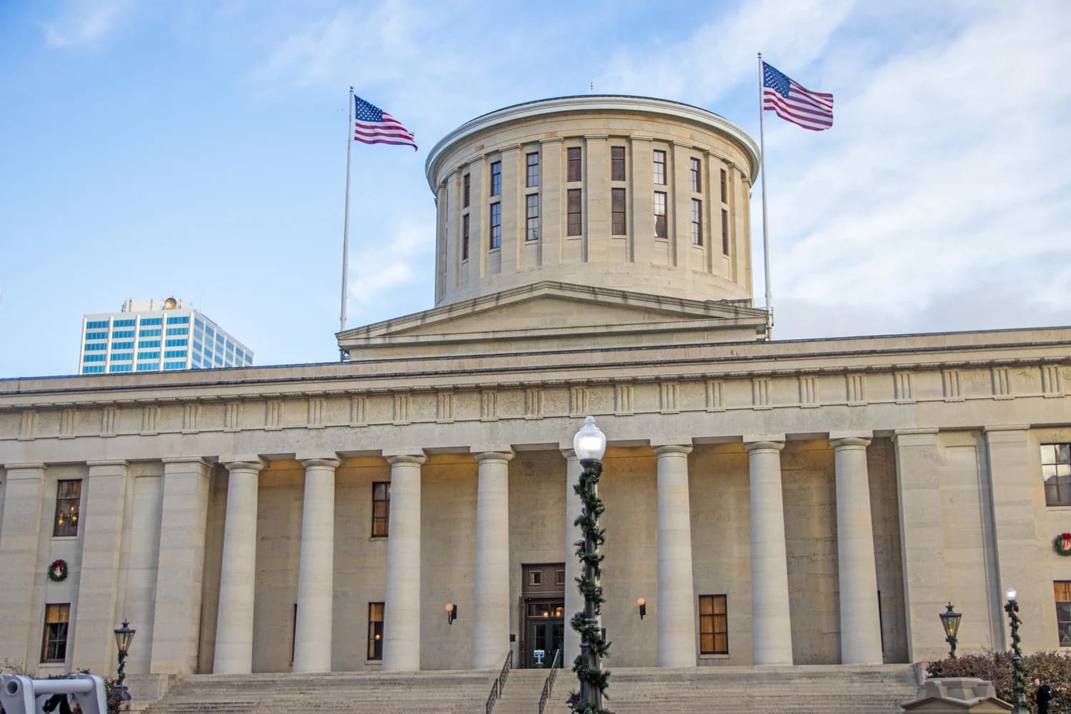 A large, light-colored stone building with a prominent dome and classical columns is shown under a partly cloudy sky. Two American flags fly from flagpoles on either side of the dome, and a modern high-rise building is visible in the background to the left.