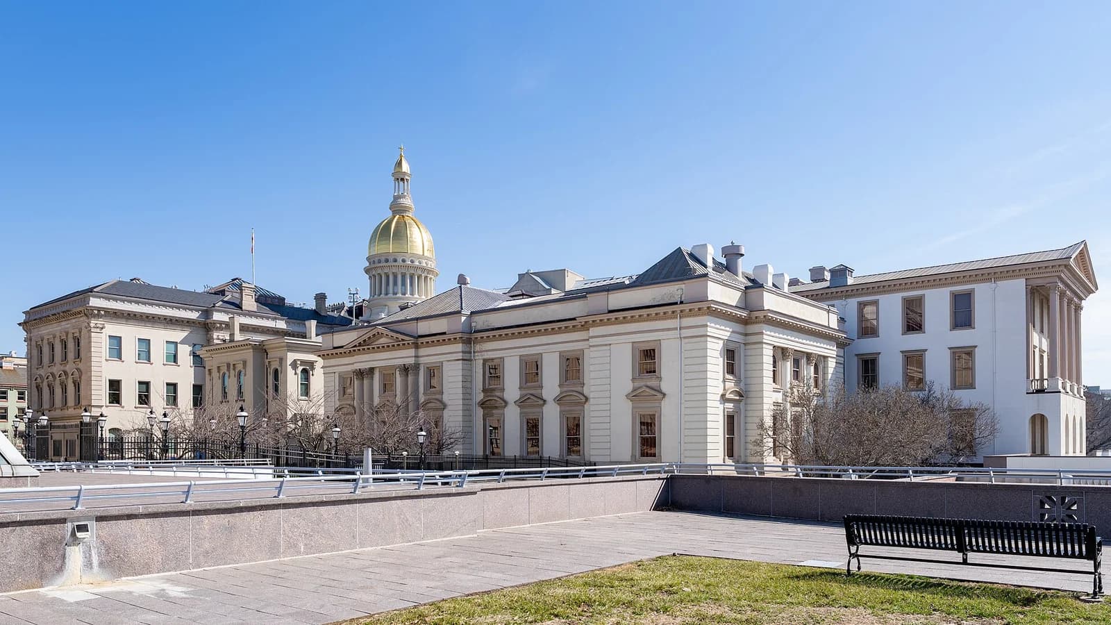 A large, light-colored institutional building with a prominent golden dome is visible under a clear blue sky. The building features classical architectural elements, including columns and ornate window details, and is viewed from a slightly elevated perspective with a paved foreground and a black park bench.