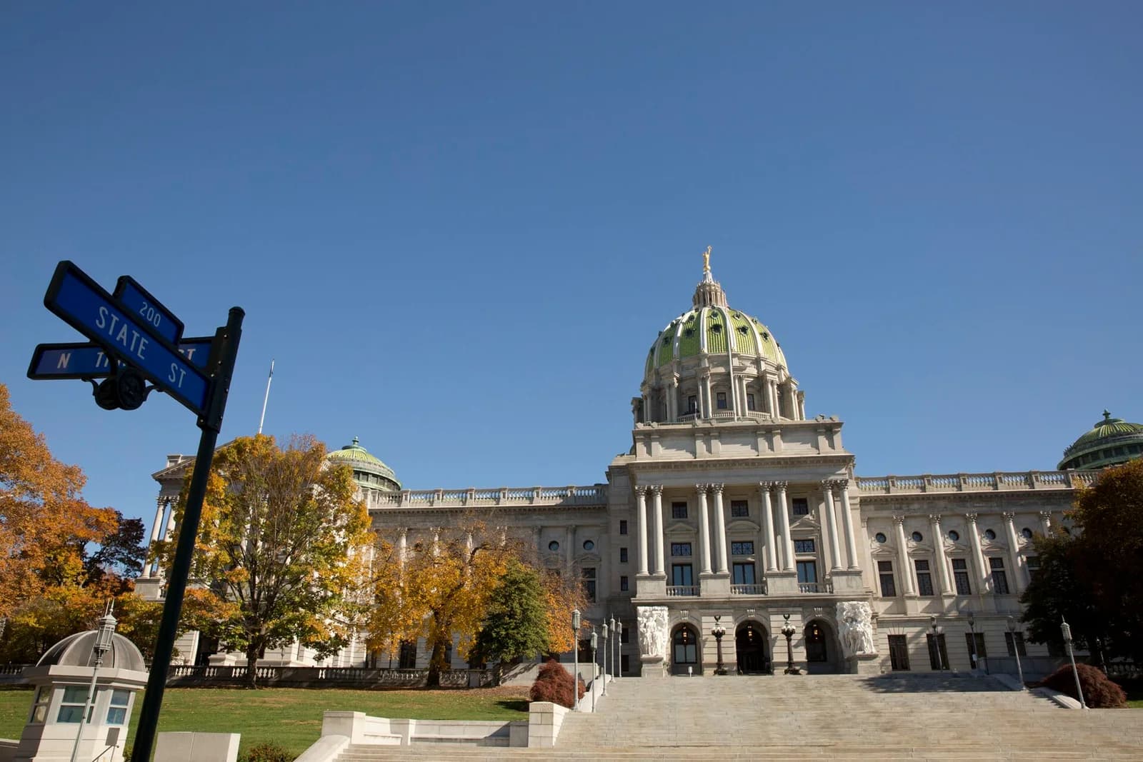 A grand stone building with a large green dome and columns stands under a clear blue sky. A street sign for "State St" is visible in the foreground to the left, with autumn trees surrounding the building.