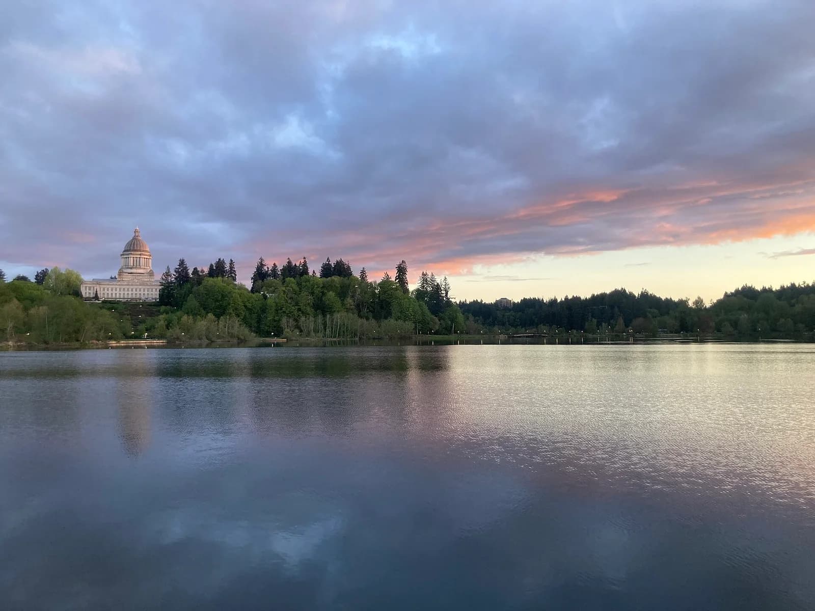 A large domed building with columns sits atop a hill surrounded by trees, overlooking a calm body of water. The sky above is cloudy with hints of pink and orange from the sunset.