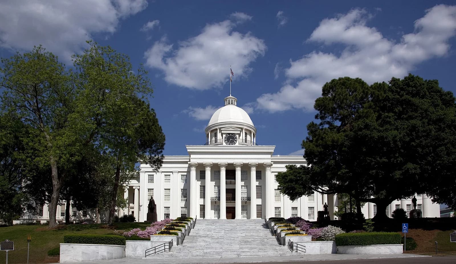 A grand white neoclassical building with a central dome and clock tower stands under a blue sky with white clouds. A wide set of steps leads up to the building, flanked by manicured hedges, flowering plants, and large green trees on either side.