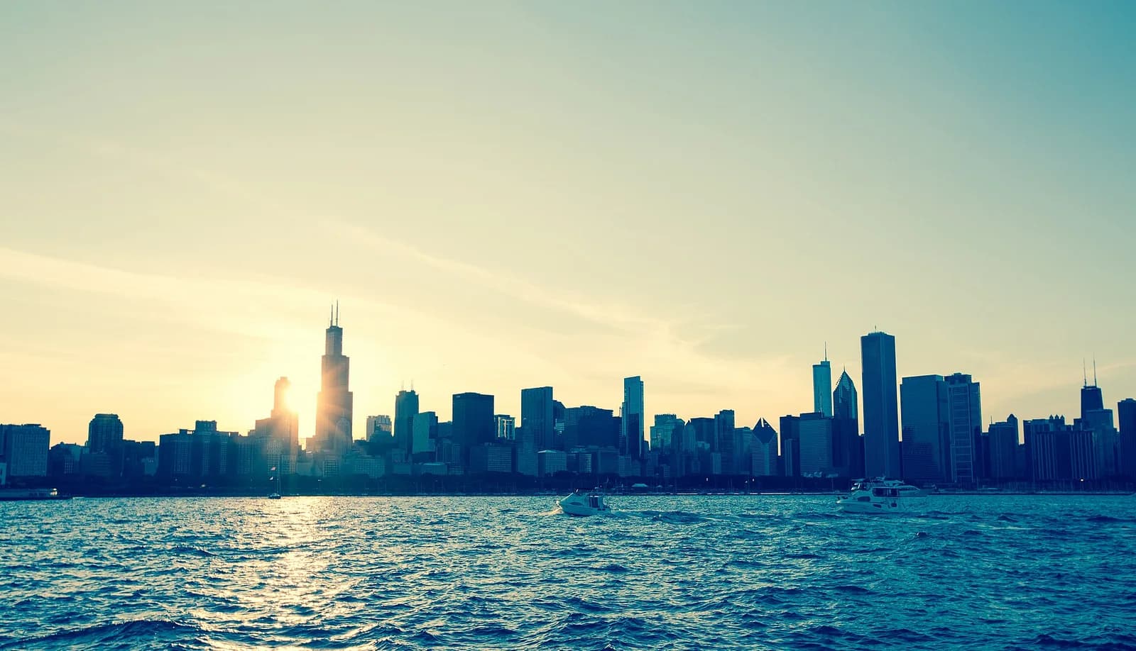 A wide shot of a city skyline at sunset, with the sun setting behind a prominent skyscraper. The foreground shows a body of water with several small boats.