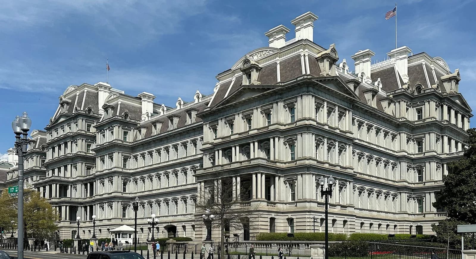 A large, ornate stone building with a mansard roof and many windows is seen from a street-level perspective under a blue sky. An American flag flies from the roof of the building on the right.