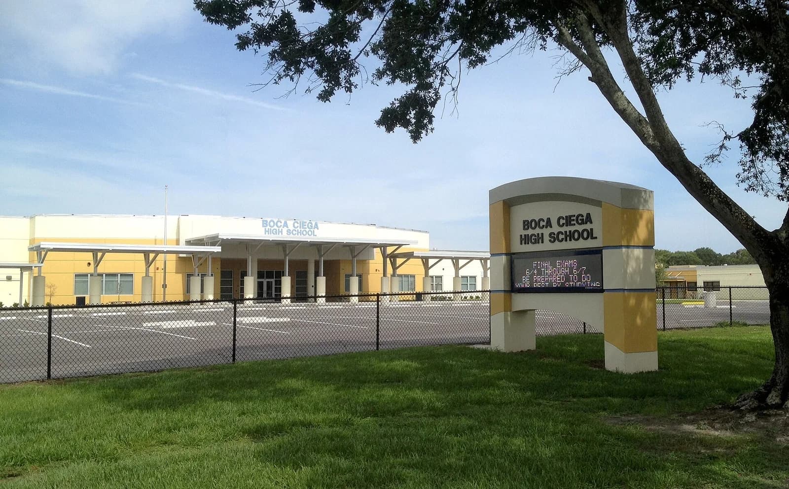 A modern school building with a yellow and white facade is visible behind a black chain-link fence and a large sign. A tree with green leaves partially obscures the right side of the image.
