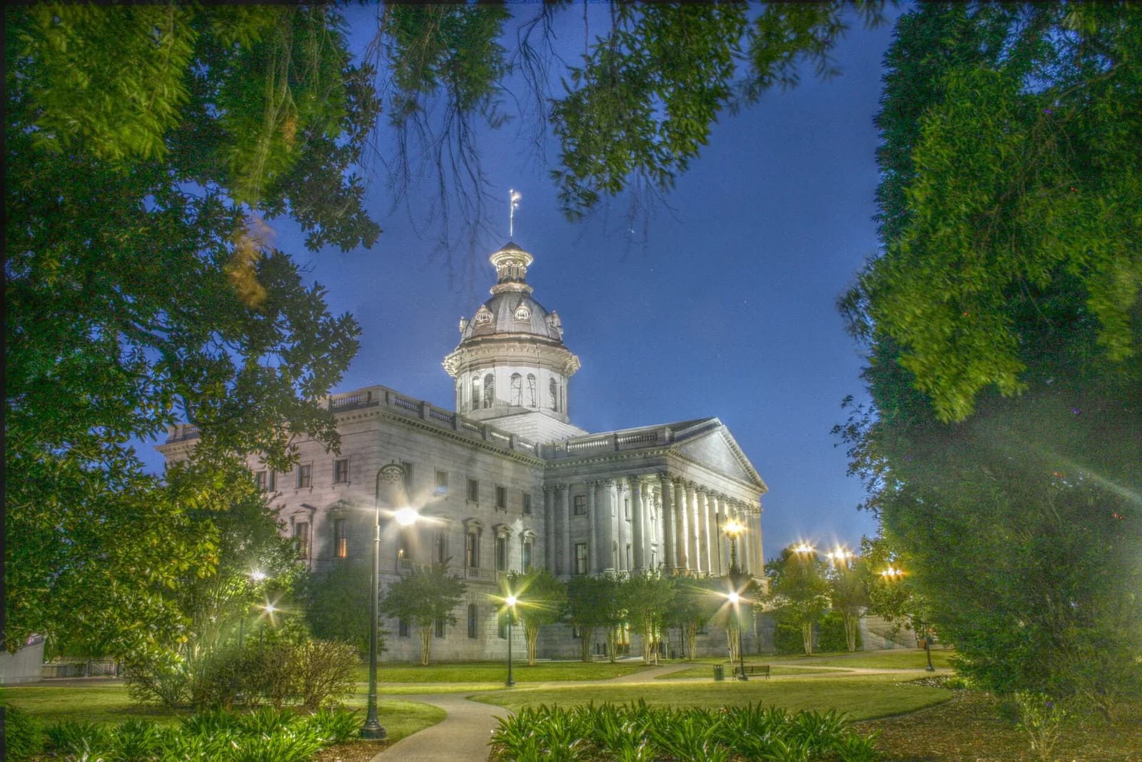 A large, classical-style building with a prominent dome and columns is illuminated at dusk, surrounded by green lawns, trees, and streetlights. A winding path leads through the foreground.