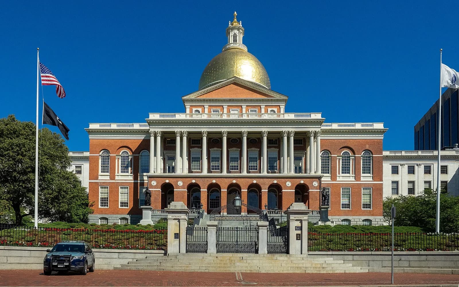 A large red brick building with a prominent gold dome and white columns stands under a clear blue sky. In the foreground, there is a black metal fence, a brick walkway, and a dark blue car parked on the left.