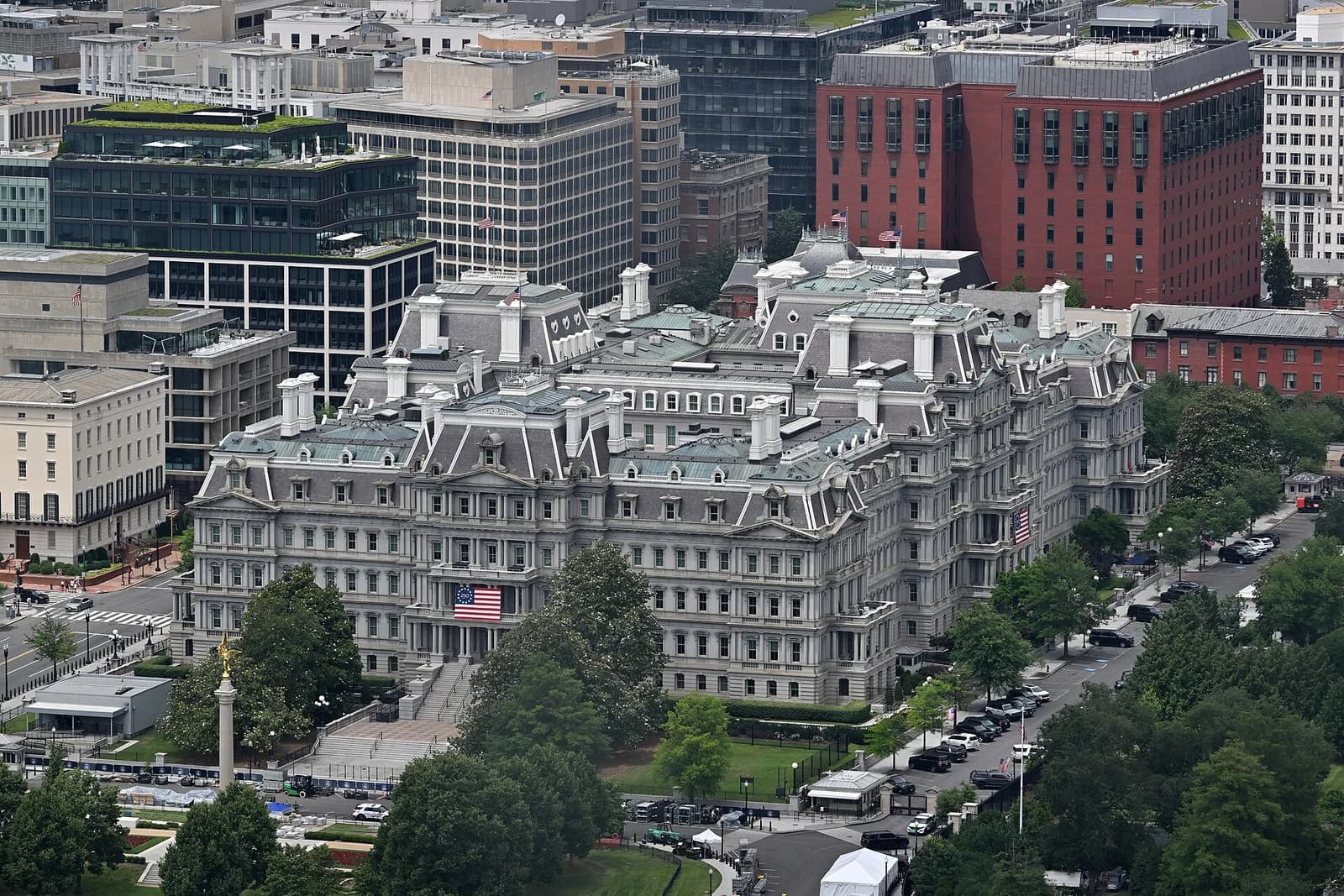 An aerial view shows a large, ornate grey stone building with a mansard roof and numerous windows, surrounded by trees and other urban buildings. An American flag hangs from the front of the building, and a street with parked cars runs alongside it.