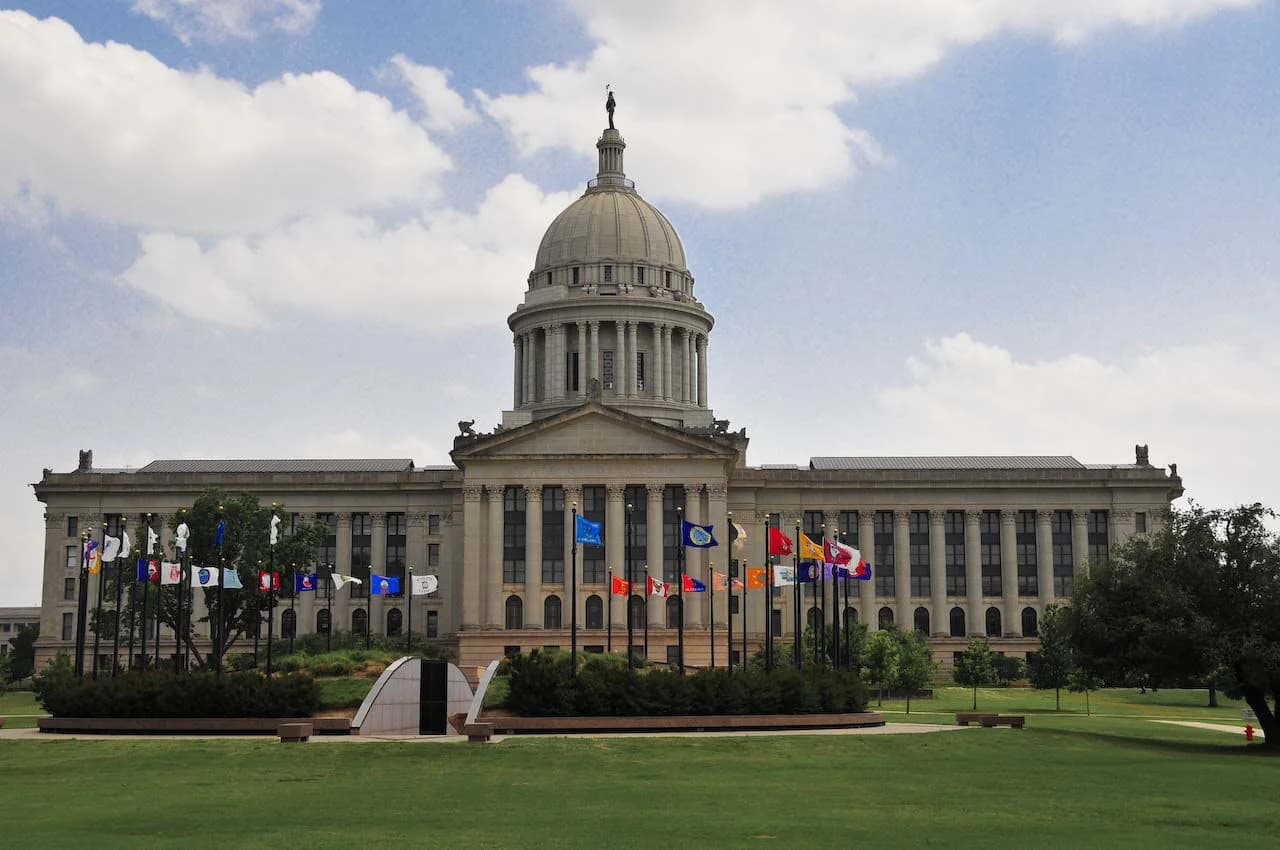 A large, classical-style building with a central dome and columns stands behind a green lawn. Numerous flags on poles are arranged in a semi-circle in front of the building.
