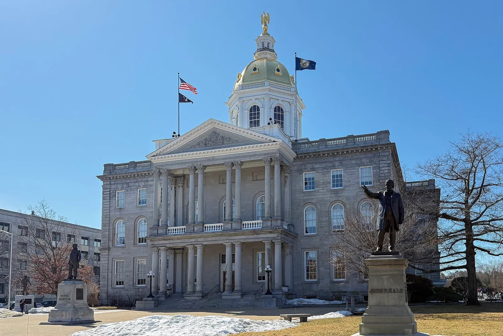 A large, light-colored stone building with a prominent portico and a golden dome topped with a statue stands under a clear blue sky. Two statues of men on pedestals are visible in the foreground, with patches of snow on the ground.