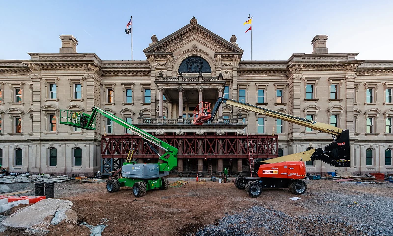 A large, ornate stone building with a central portico and flags on its roof is undergoing construction, with two boom lifts and scaffolding visible in front of it. The foreground shows a dirt construction site with debris.