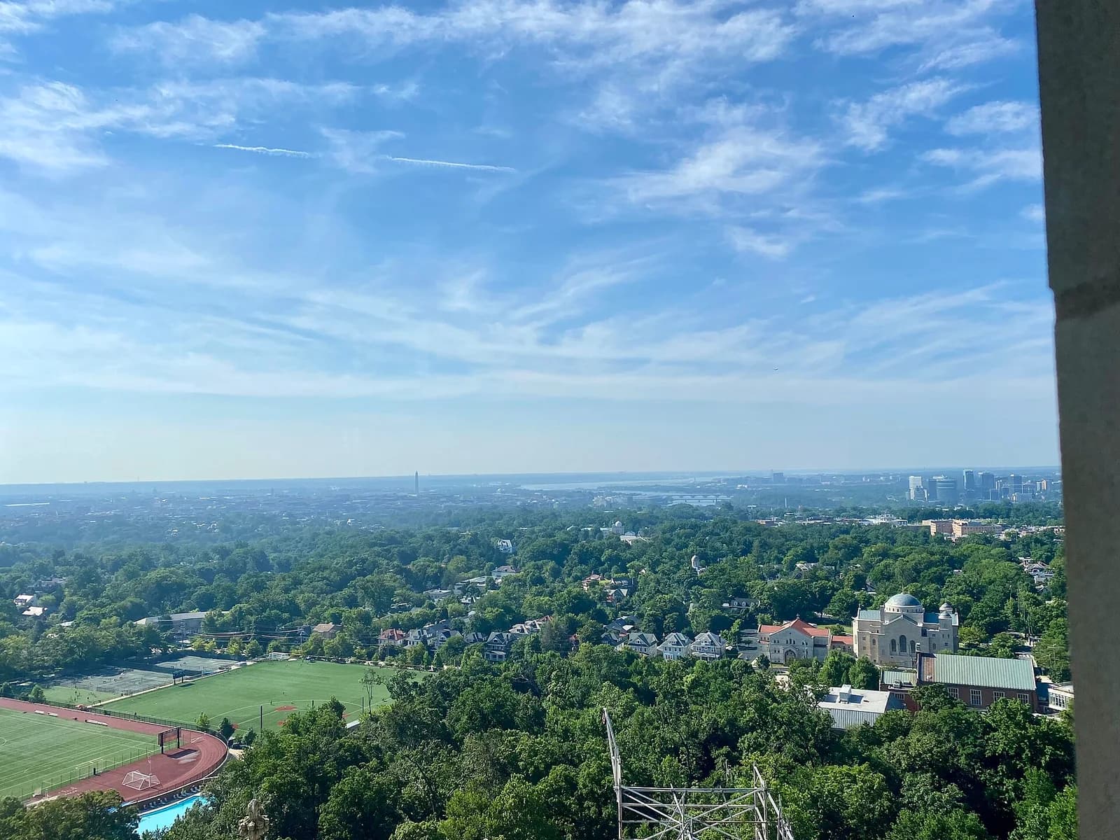 An aerial view shows a sprawling cityscape with dense green trees in the foreground, a sports field with a running track, and a large building with a dome. In the distance, a body of water and a city skyline are visible under a blue sky with wispy clouds.