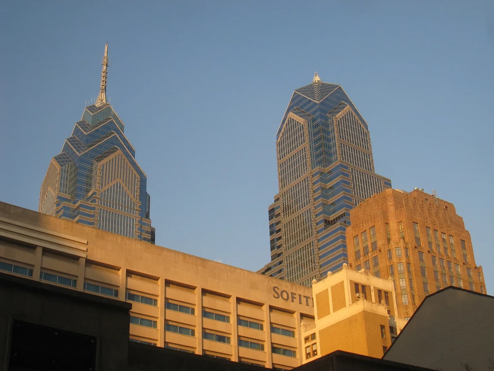 Two modern skyscrapers with blue glass facades rise above several older, lower buildings, one of which has "SOFIT" visible on its side. The sky is clear blue.