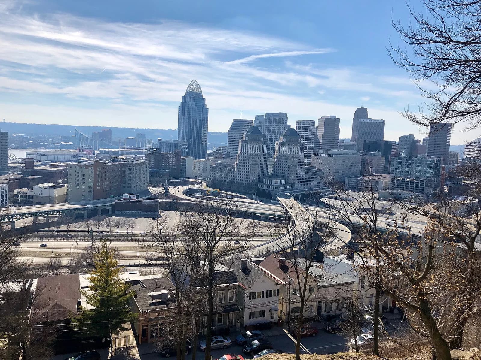 A wide view of a city skyline with numerous tall buildings under a partly cloudy sky. In the foreground, residential buildings and bare trees are visible, with highways and bridges leading into the city center.