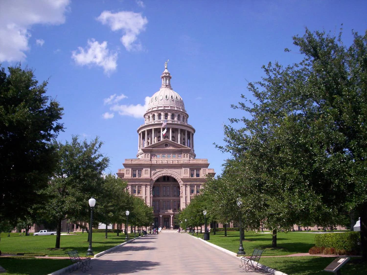 A grand, light-colored domed building with columns and arched entrances stands at the end of a wide paved path, flanked by green lawns and rows of trees. The sky is blue with scattered white clouds.