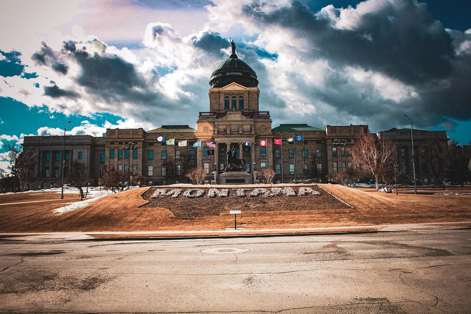 A large, classical-style building with a prominent dome and a statue in front is visible under a dramatic sky with dark clouds and patches of blue. The word "MONTANA" is spelled out in white letters on a grassy slope in the foreground.