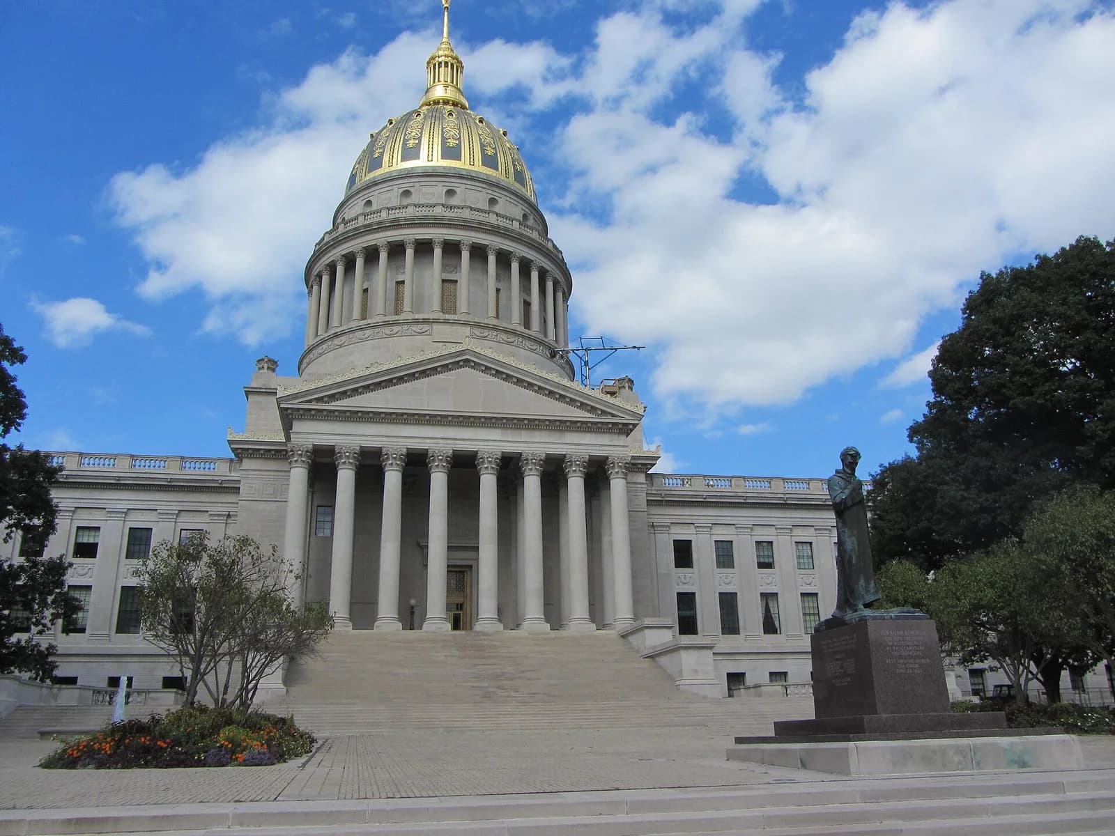 A large, light-colored stone building with a prominent golden dome and a grand staircase leading to its entrance. A bronze statue stands on a pedestal to the right of the stairs, with trees and a partly cloudy blue sky in the background.