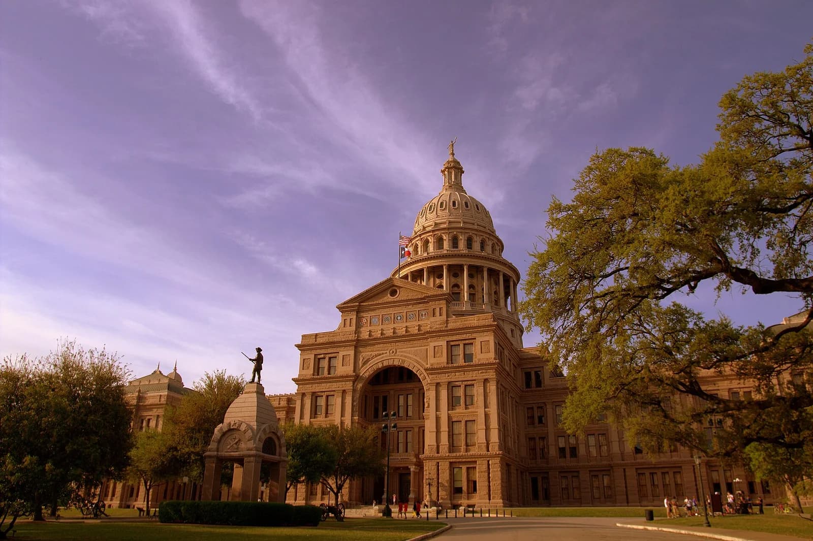 A large, ornate stone building with a prominent dome and columns stands under a partly cloudy sky. In the foreground, there is a green lawn with trees and a statue of a person holding a rifle.